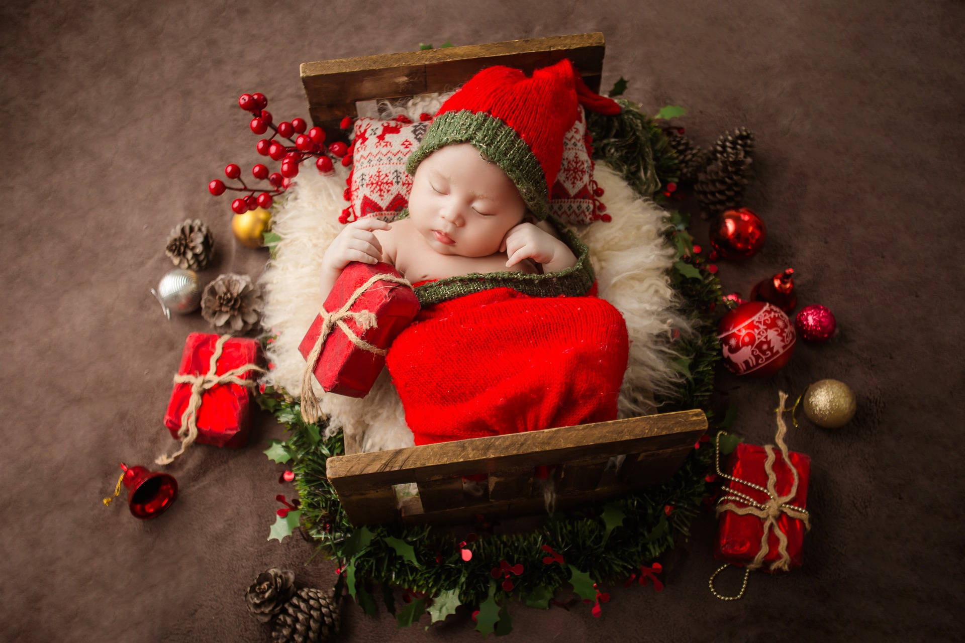 Very Cute Baby 桌布 壁紙 — 一個睡著的新生嬰兒打扮成小精靈，躺在木製雪橇裡，周圍是節日紅色禮物、松果和冬青。