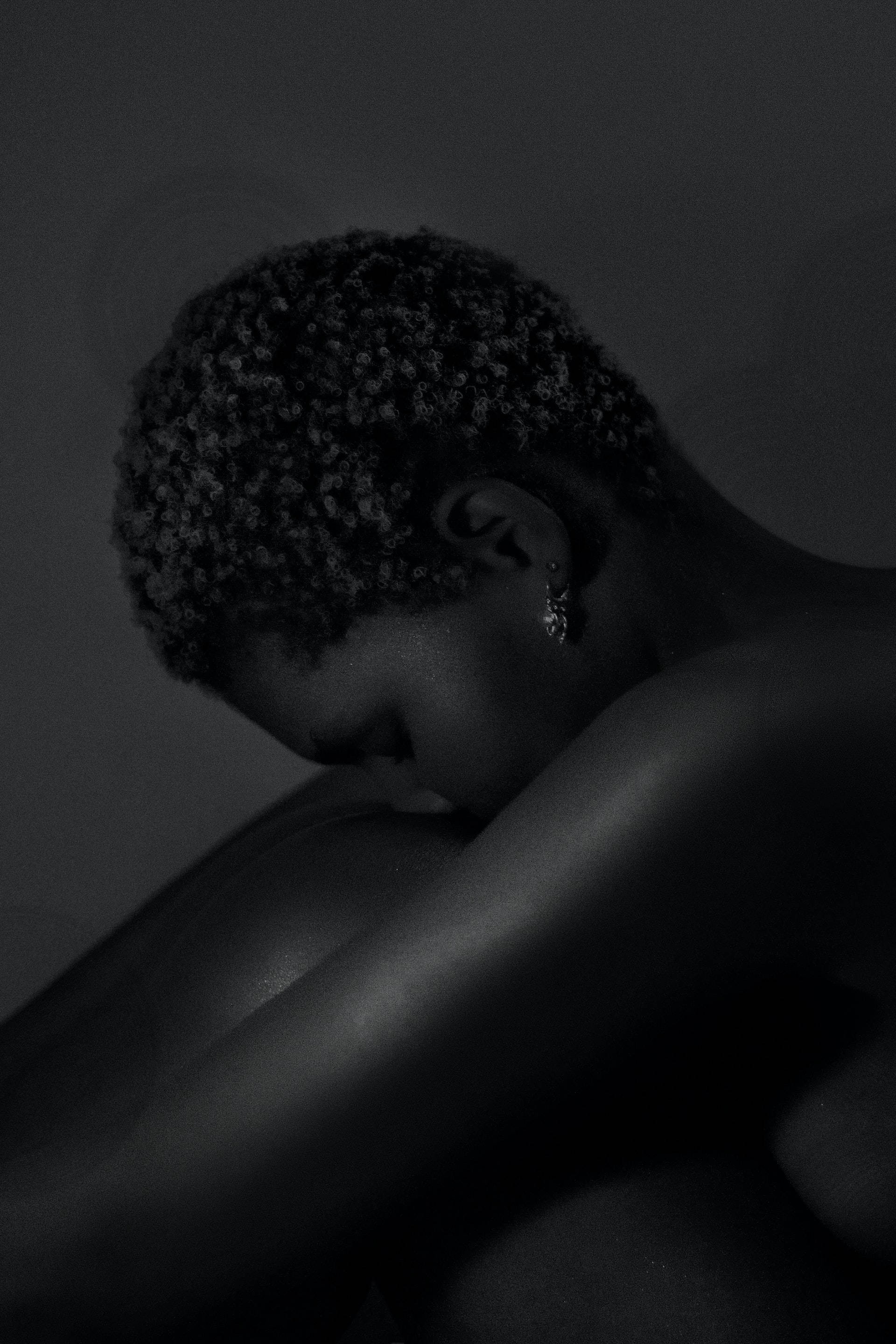 black aesthetic tumblr iPhone 桌布 壁紙 — 一個短捲髮的人在昏暗的黑白肖像中將頭靠在交叉的手臂上。