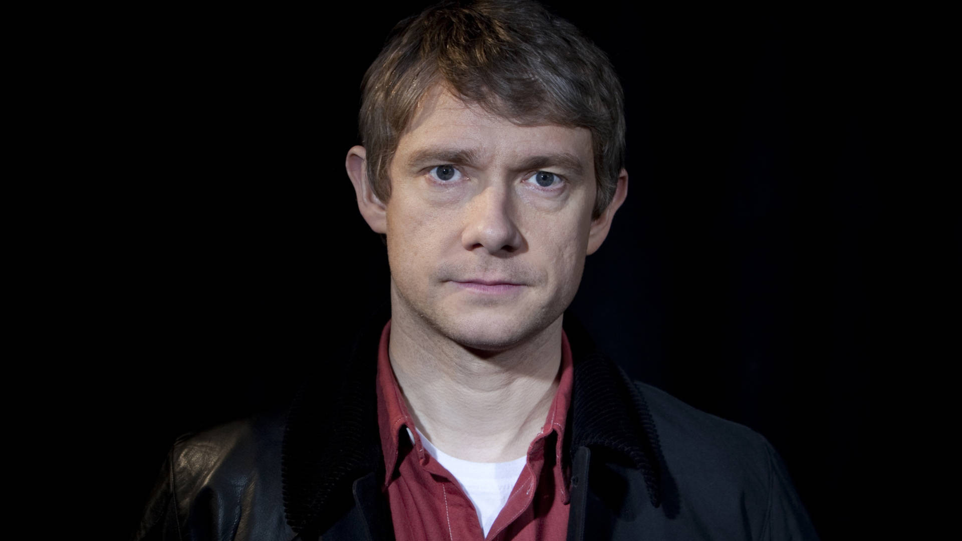Martin Freeman 桌布 壁紙 — 一個短棕髮藍眼的男人，穿著黑色外套下的紅色襯衫，在純黑色背景前直視鏡頭。