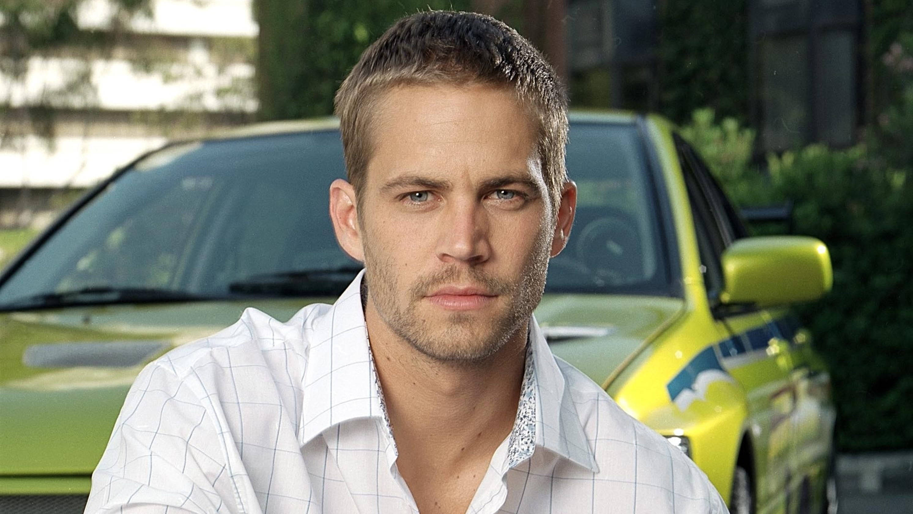 Paul Walker 桌布 壁紙 — 一個短金髮、有鬍渣的男人，穿著白色格紋襯衫，身後有一輛模糊的黃色汽車，專注地凝視著鏡頭。