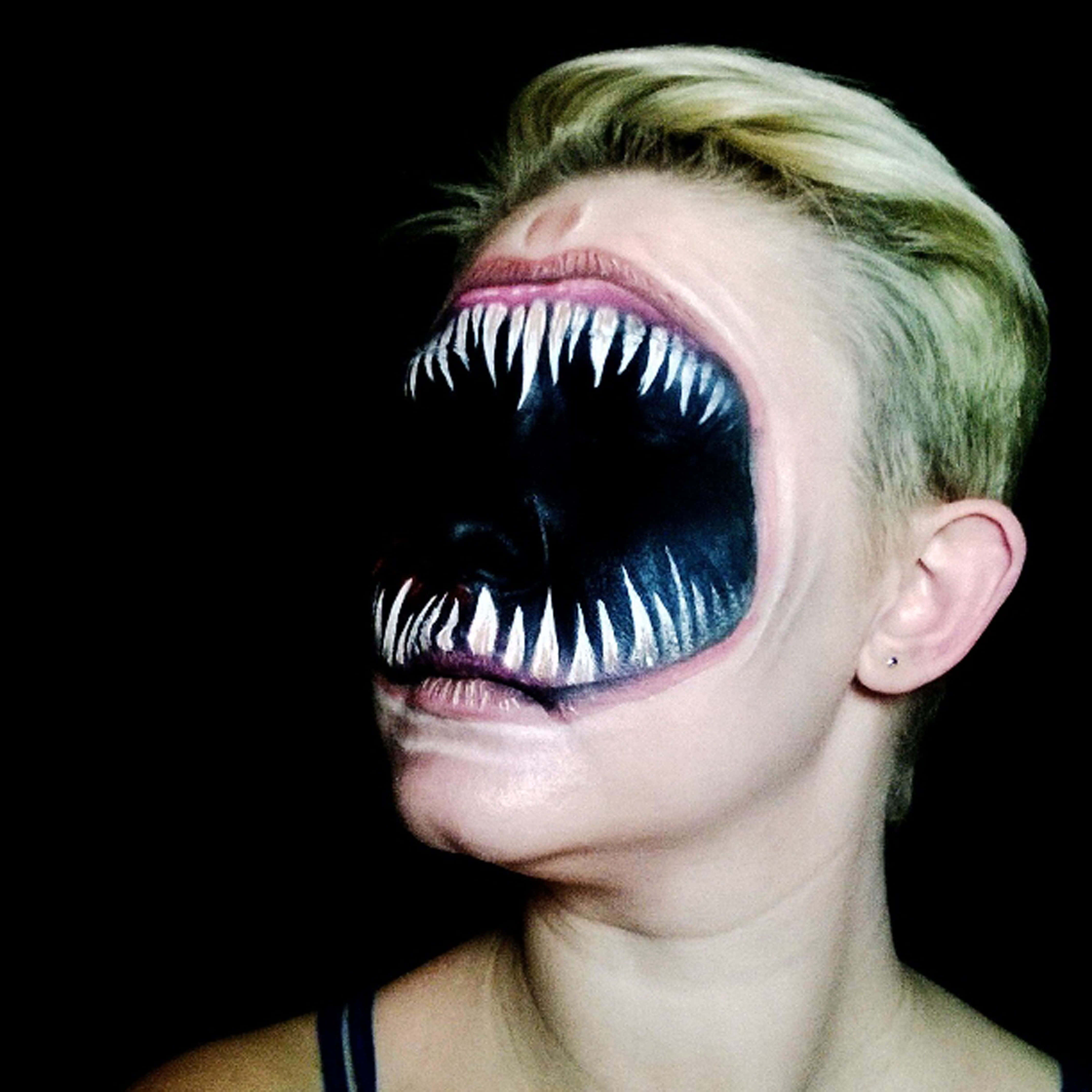 Scary Face 桌布 壁紙 — 一個短金髮的人臉上畫了一張怪異的嘴巴，有尖銳的白色牙齒和黑色內部，背景是深色的。