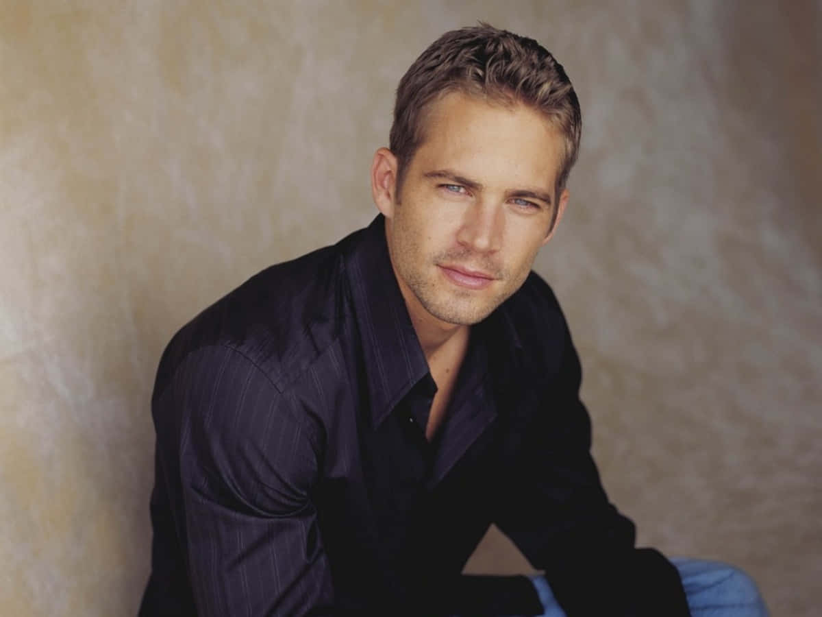 Paul Walker 桌布 壁紙 — 一個短金髮藍眼的男人，穿著深色細條紋襯衫，靠在有紋理的米色牆上。