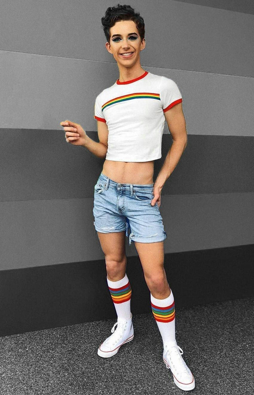 twink 桌布 壁紙 — 一個短黑髮、化著大膽妝容的人，穿著彩虹條紋短版上衣、牛仔短褲和彩虹襪子，在灰色牆壁前微笑。