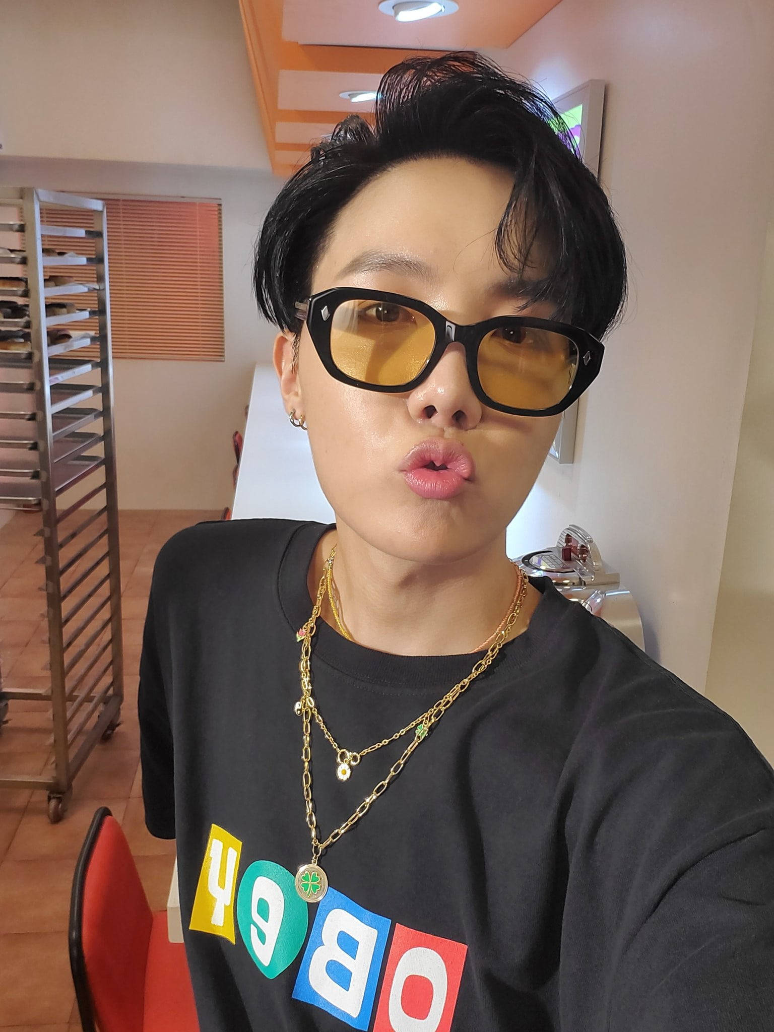 jhope 桌布 壁紙 — 一個短黑髮、戴黃色太陽眼鏡的人，在廚房穿著黑色圖案T恤，做出俏皮的嘟嘴表情。
