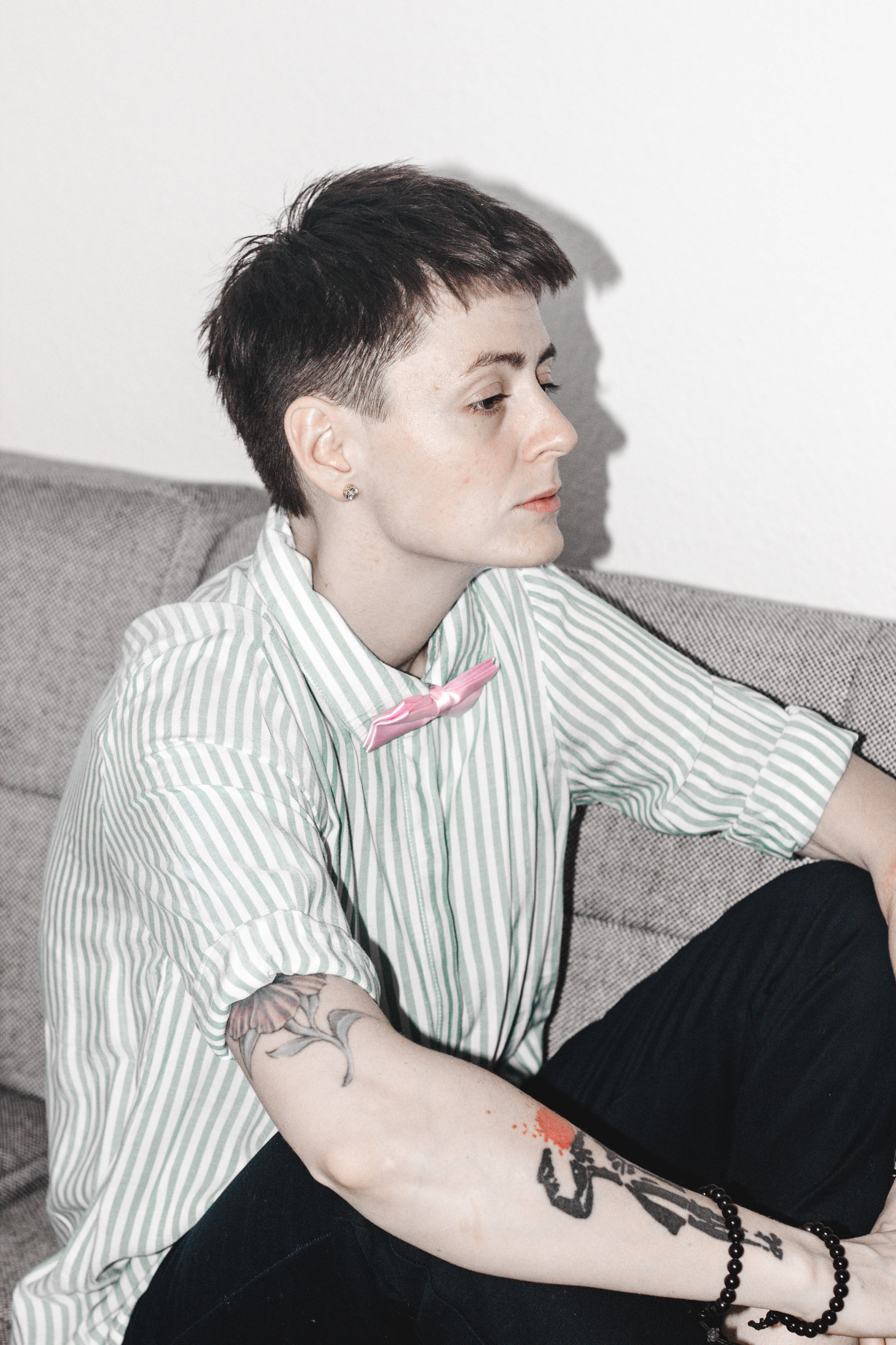 Tomboy 桌布 壁紙 — 一個短黑髮、有刺青的人坐在灰色沙發上，穿著條紋襯衫和粉紅色蝴蝶結。