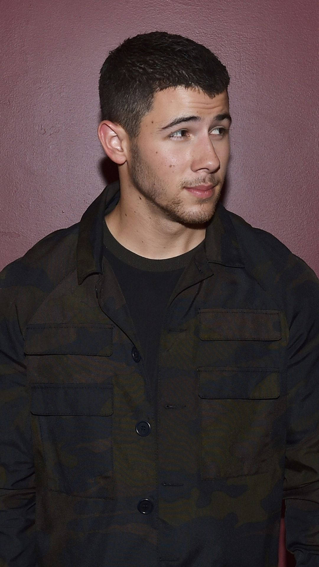 Nick Jonas 桌布 壁紙 — 一個短黑髮、留鬍渣的男人，穿著迷彩外套，靠著柔和的粉紅色牆壁。