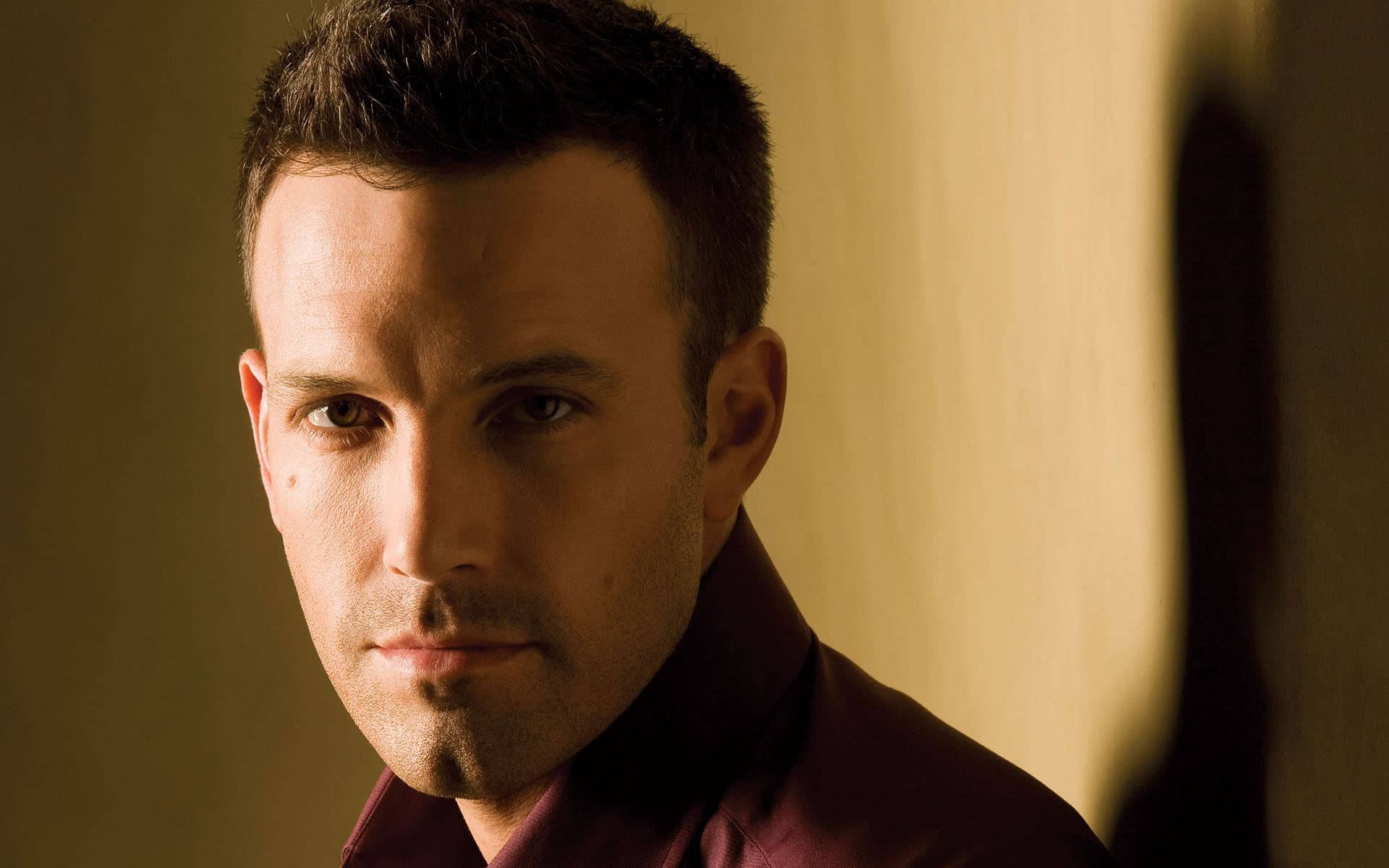 Ben Affleck 桌布 壁紙 — 一個短黑髮、身穿栗色襯衫的男人，在溫暖的金色燈光下專注地凝視著相機。