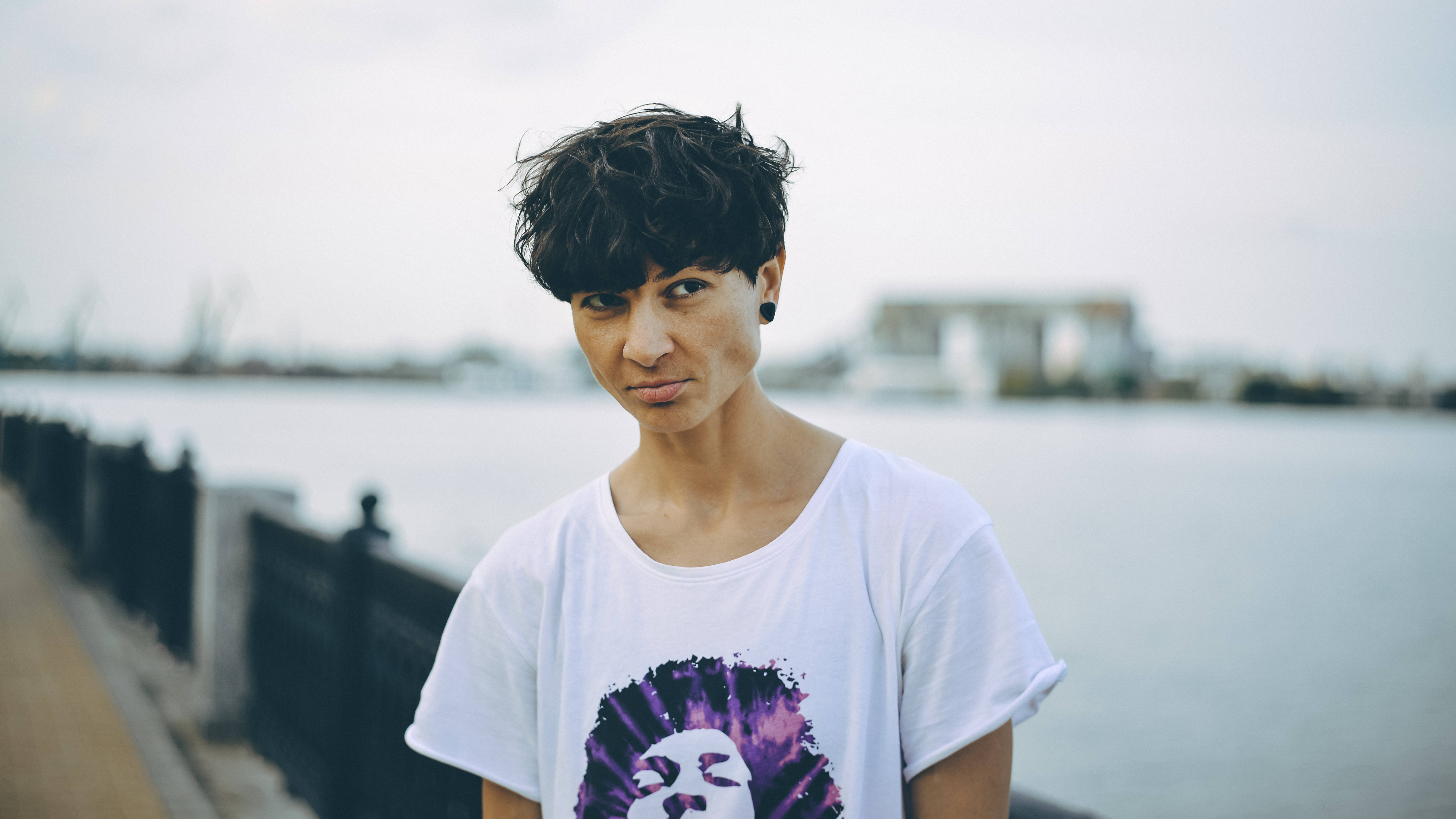 Tomboy 桌布 壁紙 — 一個短黑髮、身穿白色T恤的人站在多雲天空下的水邊欄杆旁。