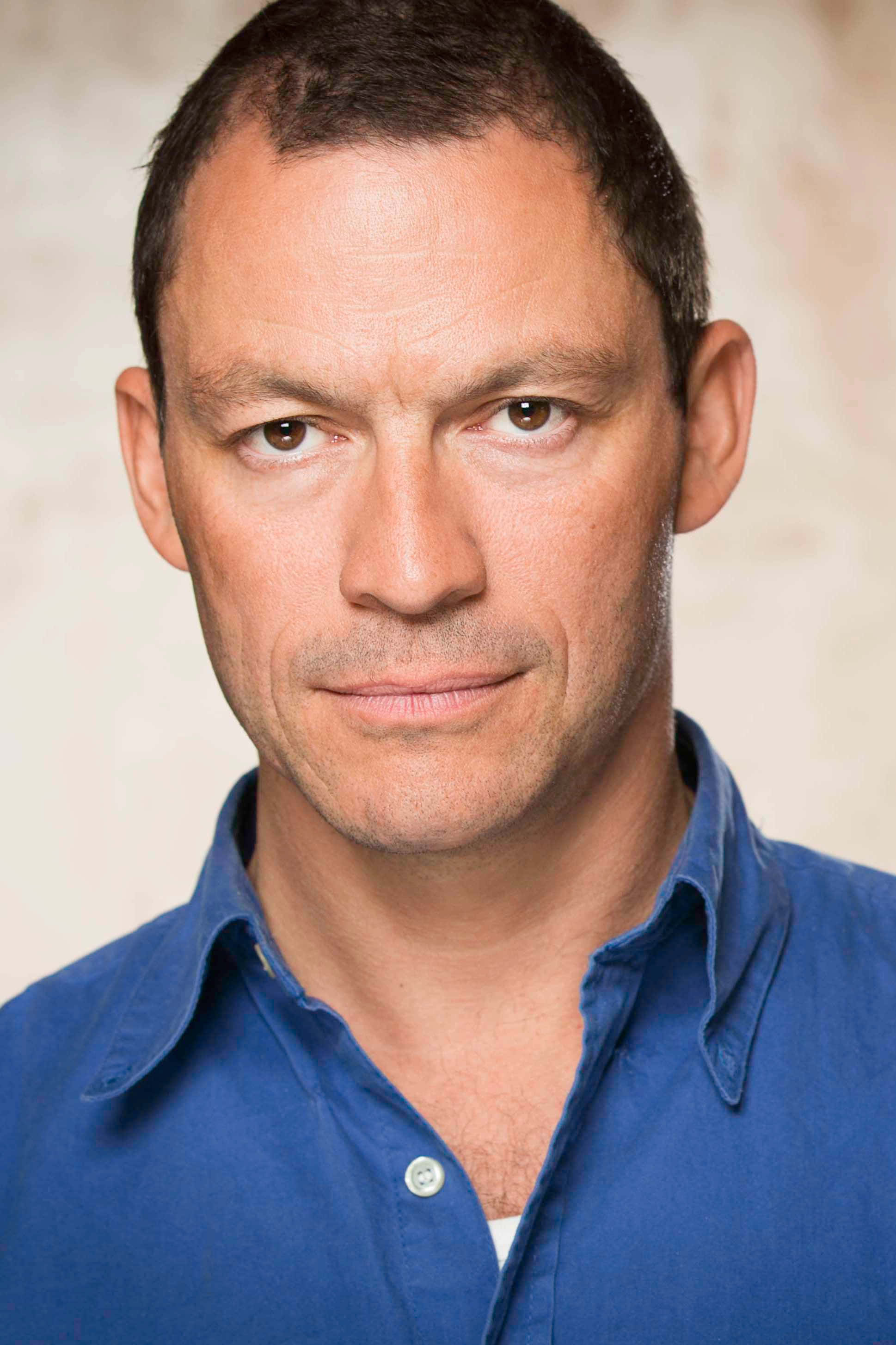 Dominic West 桌布 壁紙 — 一個短黑髮男人穿著藍色鈕扣襯衫，在柔和米色背景前的特寫肖像。