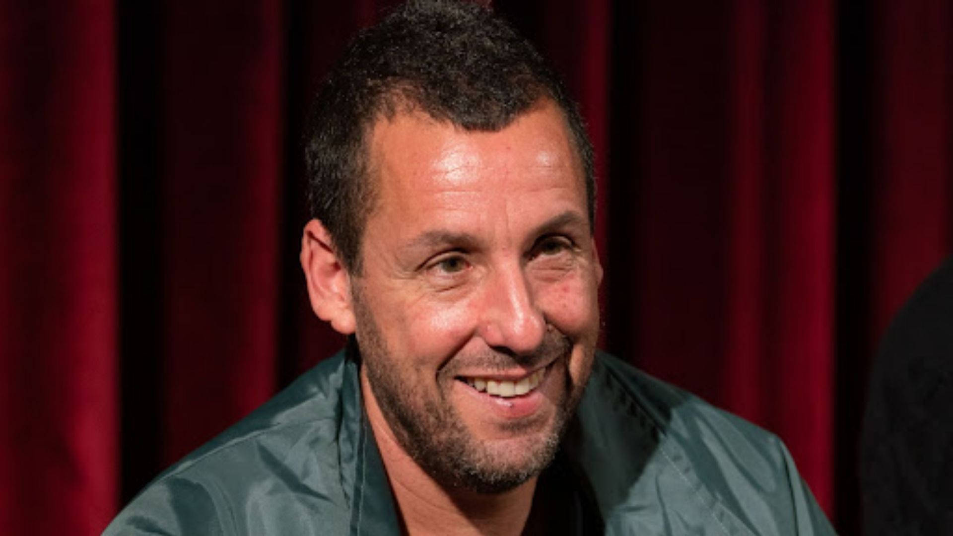 Adam Sandler 桌布 壁紙 — 一個短黑髮留鬍子的男人，在深紅色窗簾背景下溫暖地微笑。