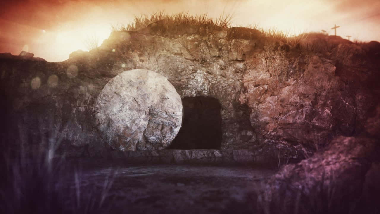 Holy Saturday 桌布 壁紙 — 一個石墓，蓋子被滾到一旁，位於岩石遍地的田野中，上方是溫暖燦爛的日落天空。