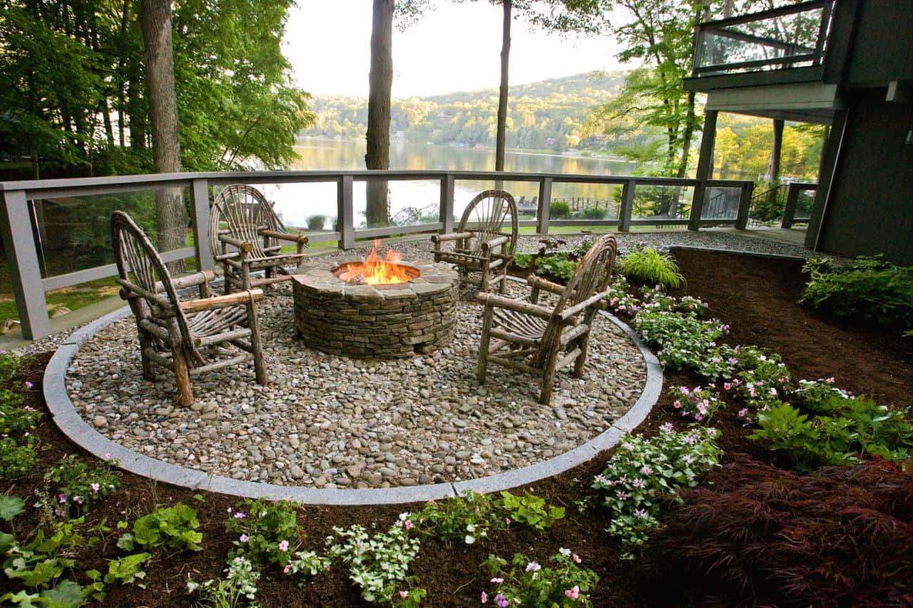 Garden Design 桌布 壁紙 — 一個石頭火坑，閃爍的火焰位於碎石圓圈內，周圍是質樸的木椅和盛開的花朵，位於一個現代的甲板上，俯瞰著寧靜的湖泊。