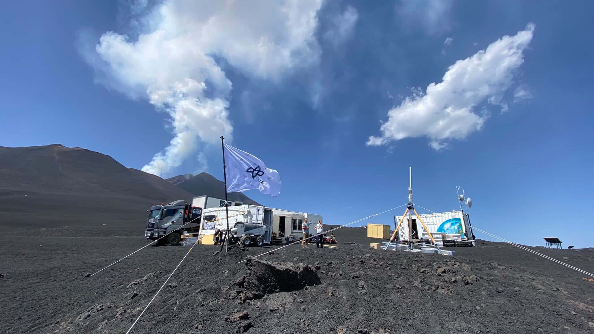 Trucks 桌布 壁紙 — 一個研究站位於黑色火山景觀上，天空明亮湛藍，有白雲和從遠處山脈升起的煙羽。
