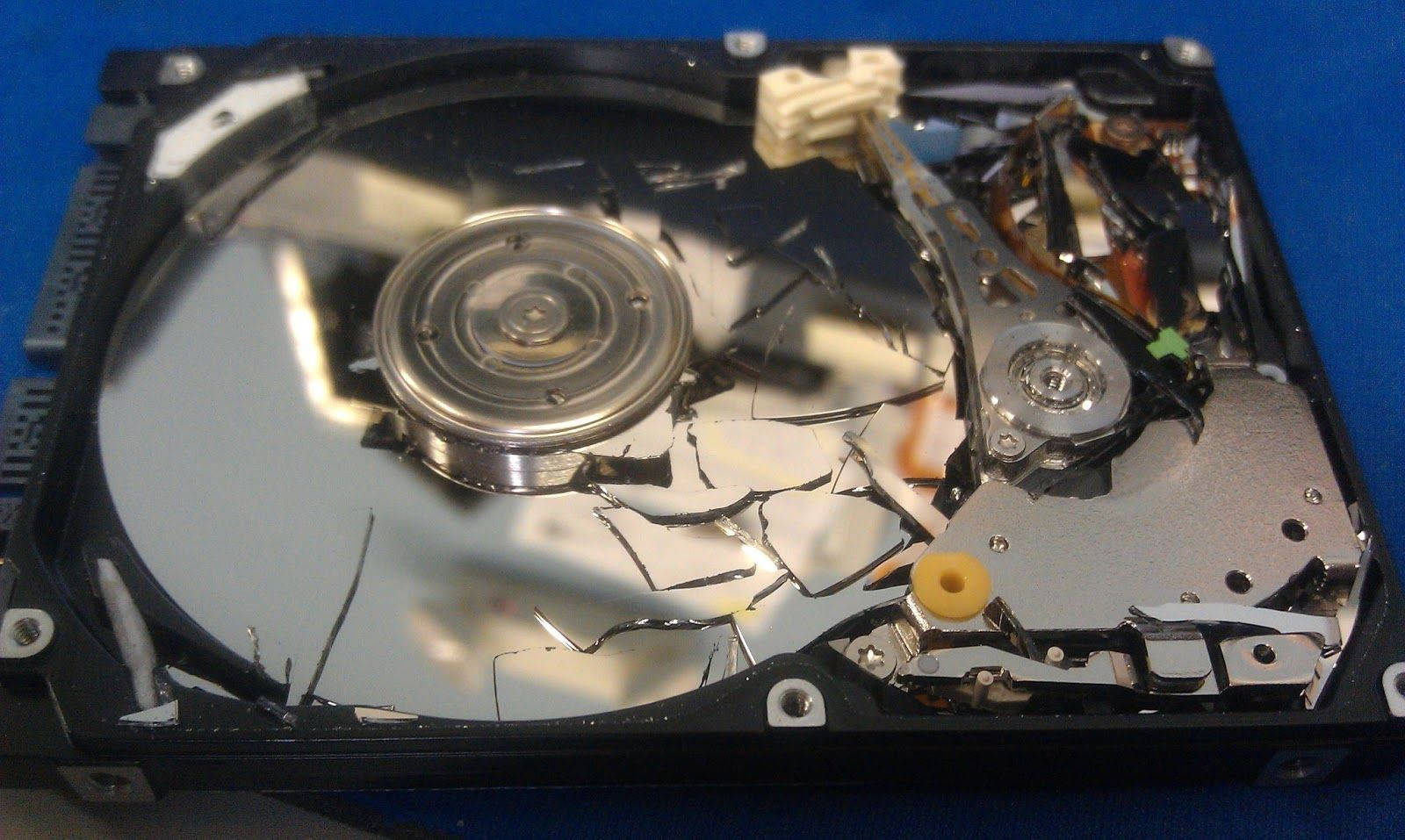 Hard Drive 桌布 壁紙 — 一個破碎的硬碟，帶有損壞的碟片和暴露的內部組件，放置在藍色表面上。