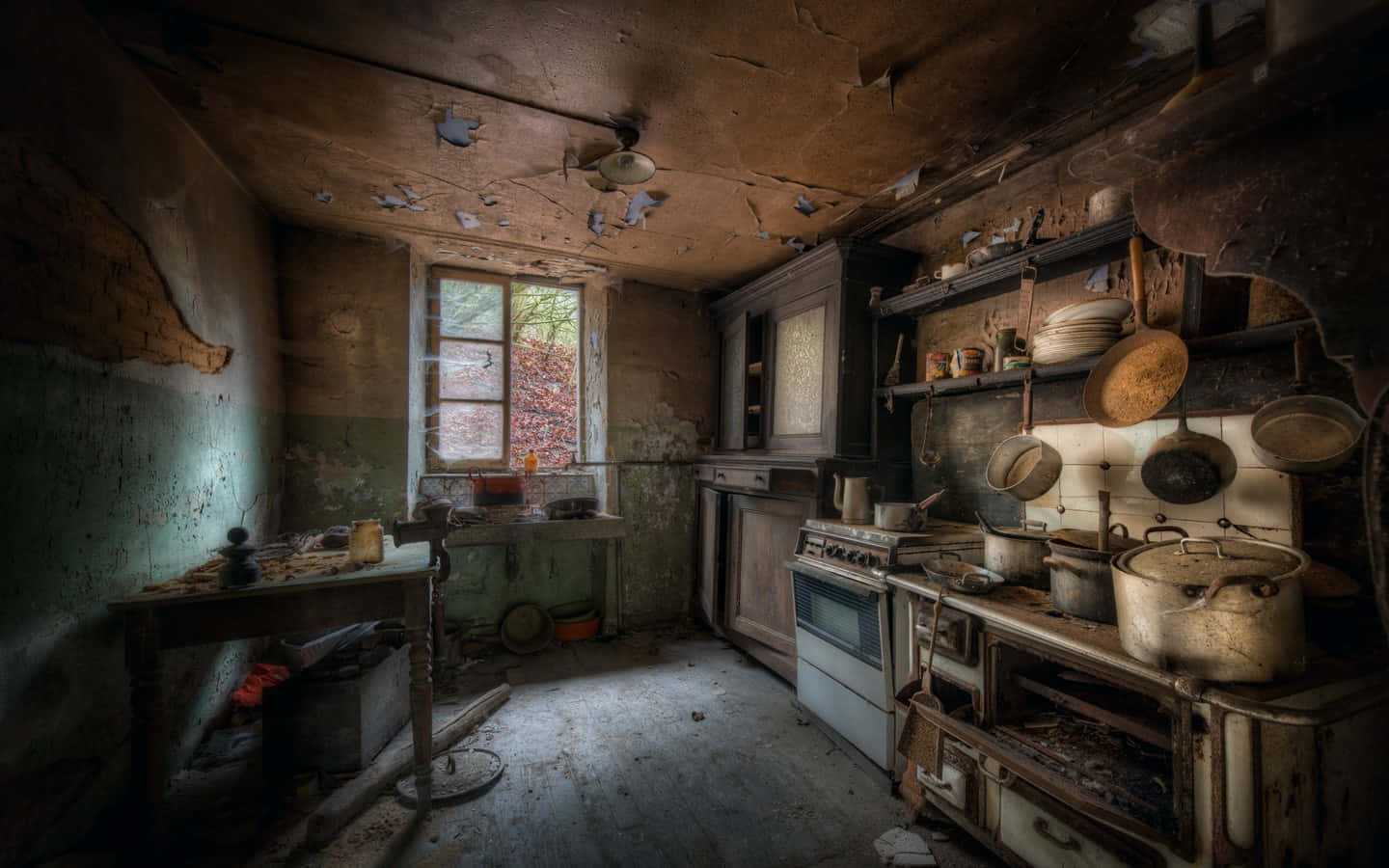 1440p Kitchen 桌布 壁紙 — 一個破舊的廚房，牆壁剝落，天花板破損，沐浴在單一窗戶的昏暗光線中。