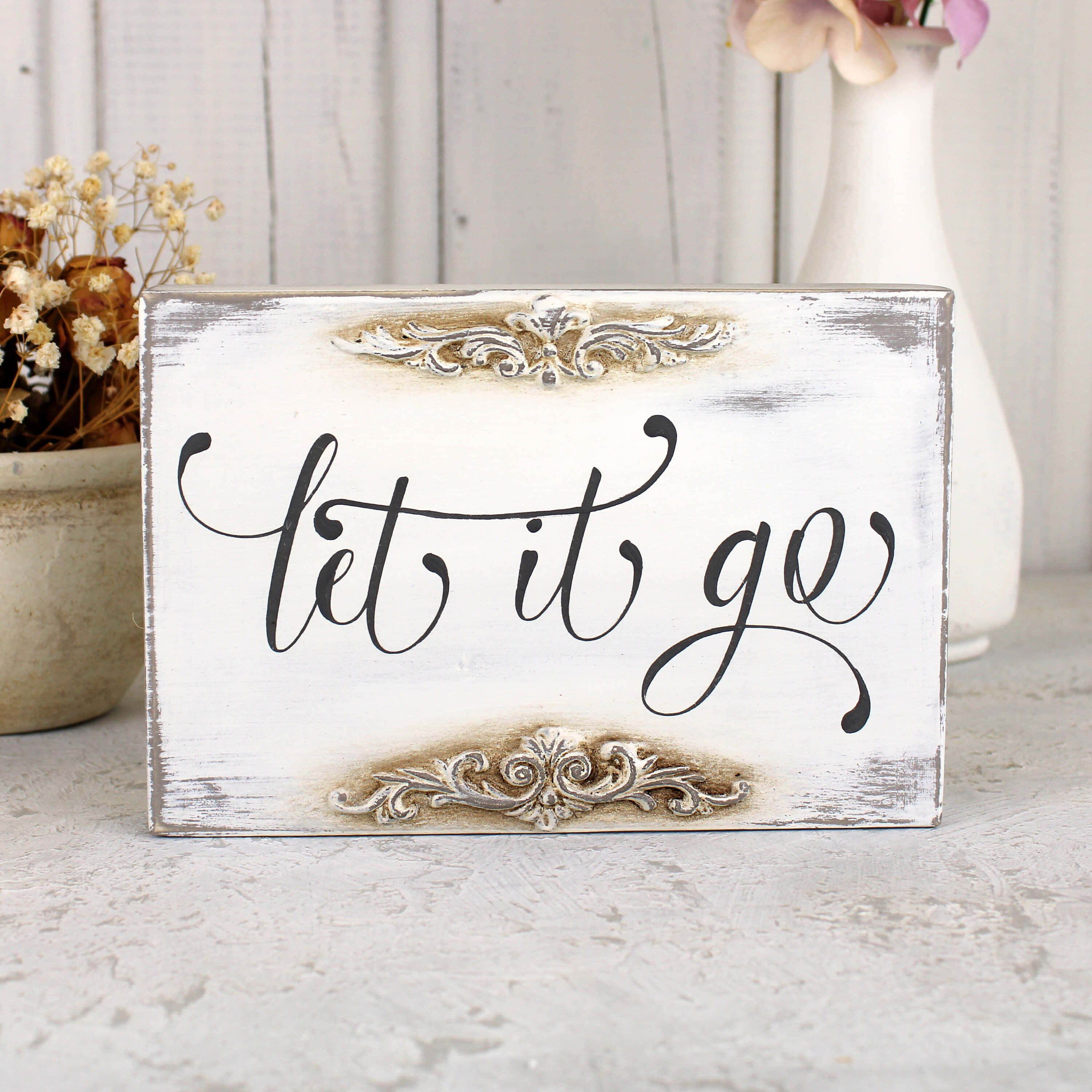 let it go 桌布 壁紙 — 一個破舊的白色木牌，上面有黑色草書文字寫著 let it go，飾有華麗的銀色裝飾，放置在帶紋理的表面上，旁邊有乾花和一個花瓶。