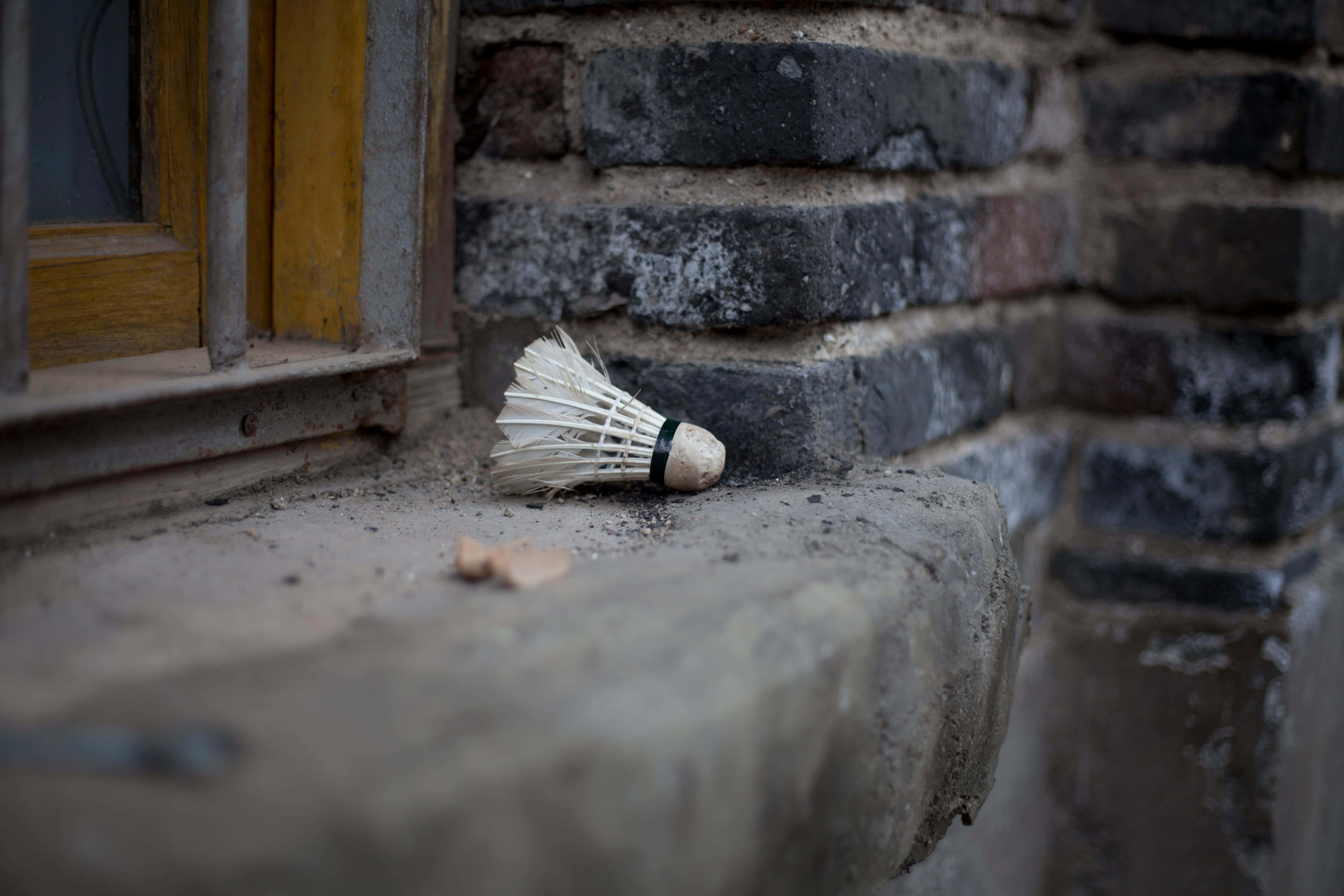 Badminton 桌布 壁紙 — 一個破舊的羽毛球在一個風化的石窗台上，旁邊是一堵鄉村風格的磚牆和一個黃色框的窗戶。
