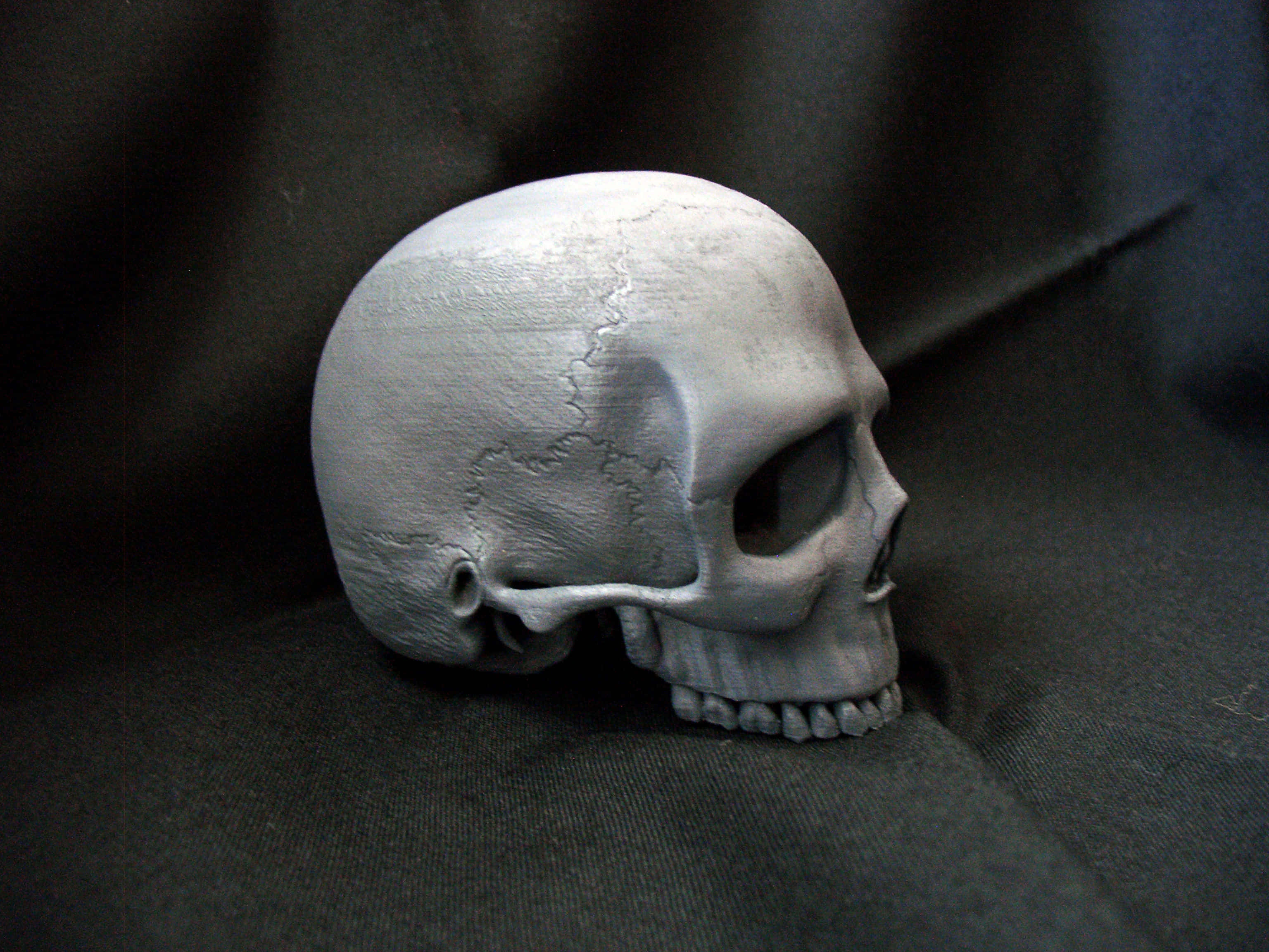 3d Skull 桌布 壁紙 — 一個破裂的灰色頭骨放在深色布料上，柔和光線投射出細微陰影。