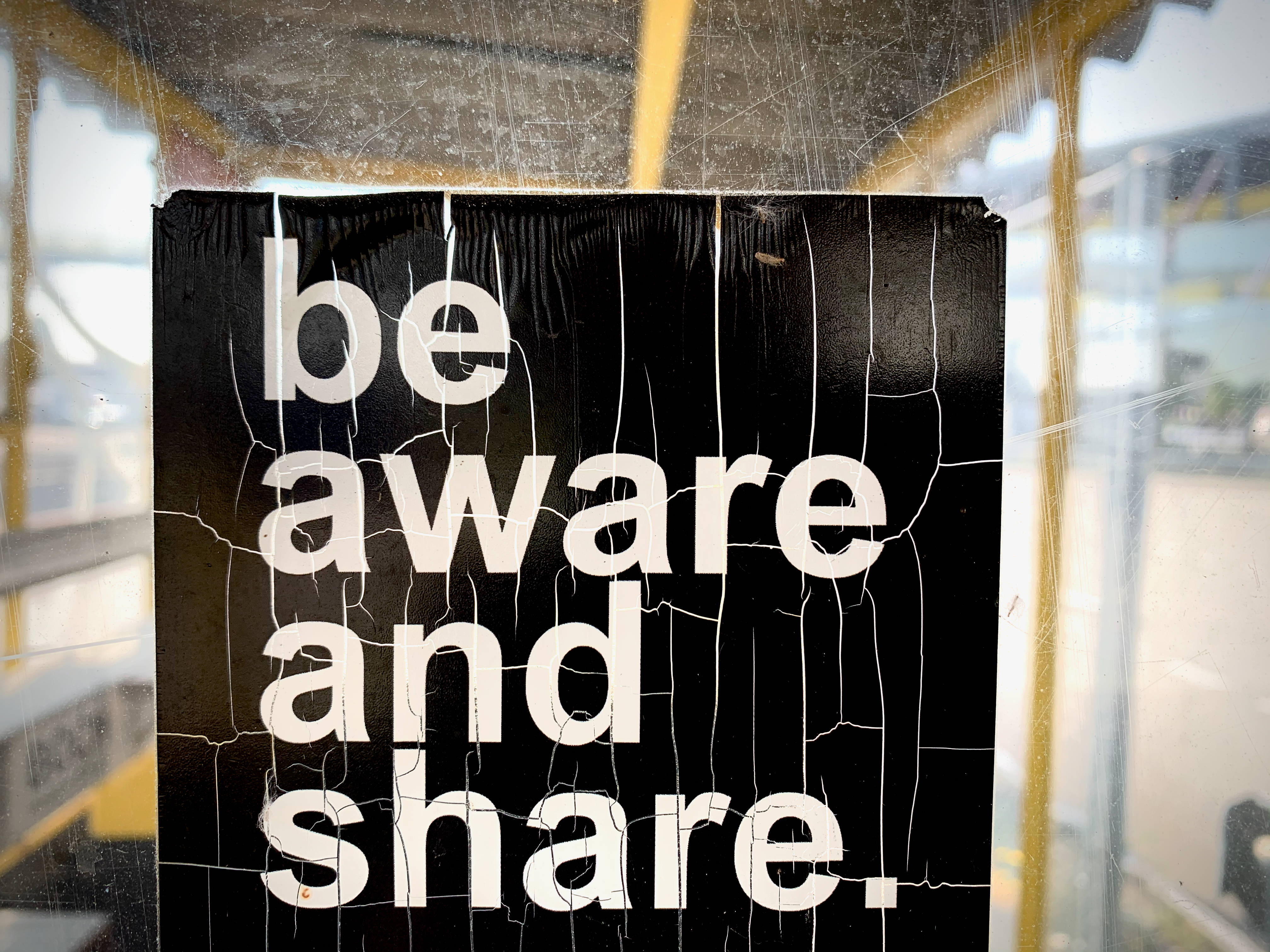 字型 桌布 壁紙 — 一個破裂的黑色標誌，上面有白色文字寫著「be aware and share」，貼在一個帶有黃色鐵欄的骯髒窗戶上。