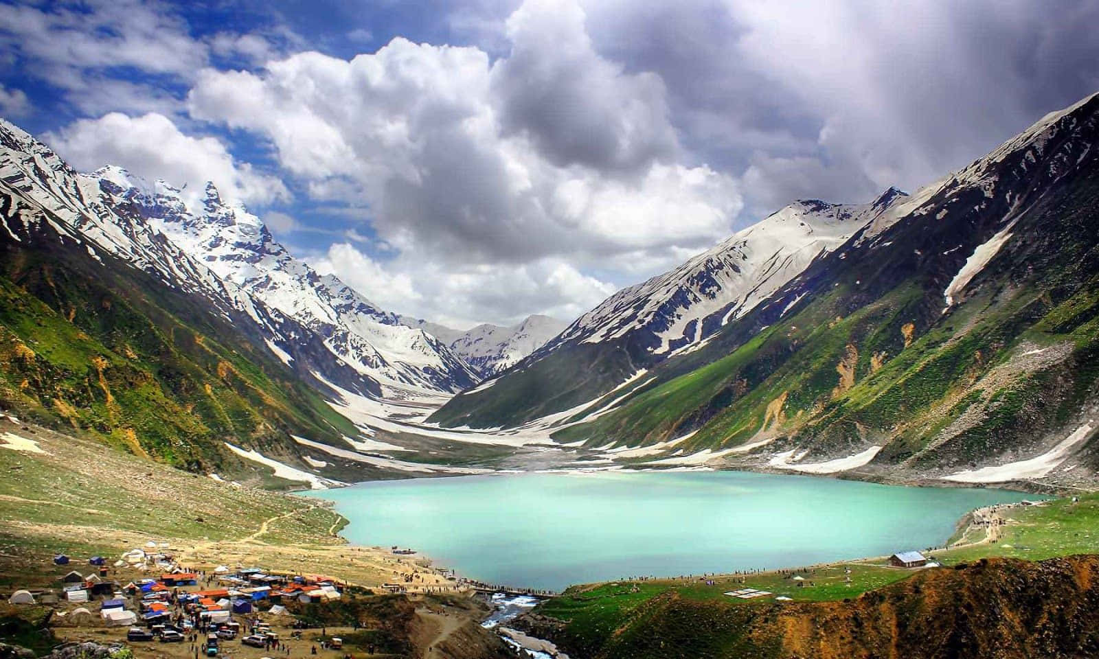 Pakistan 桌布 壁紙 — 一個碧綠色的冰川湖，坐落在山谷中，四周環繞著白雪覆蓋的山峰，上方是戲劇性的多雲天空。
