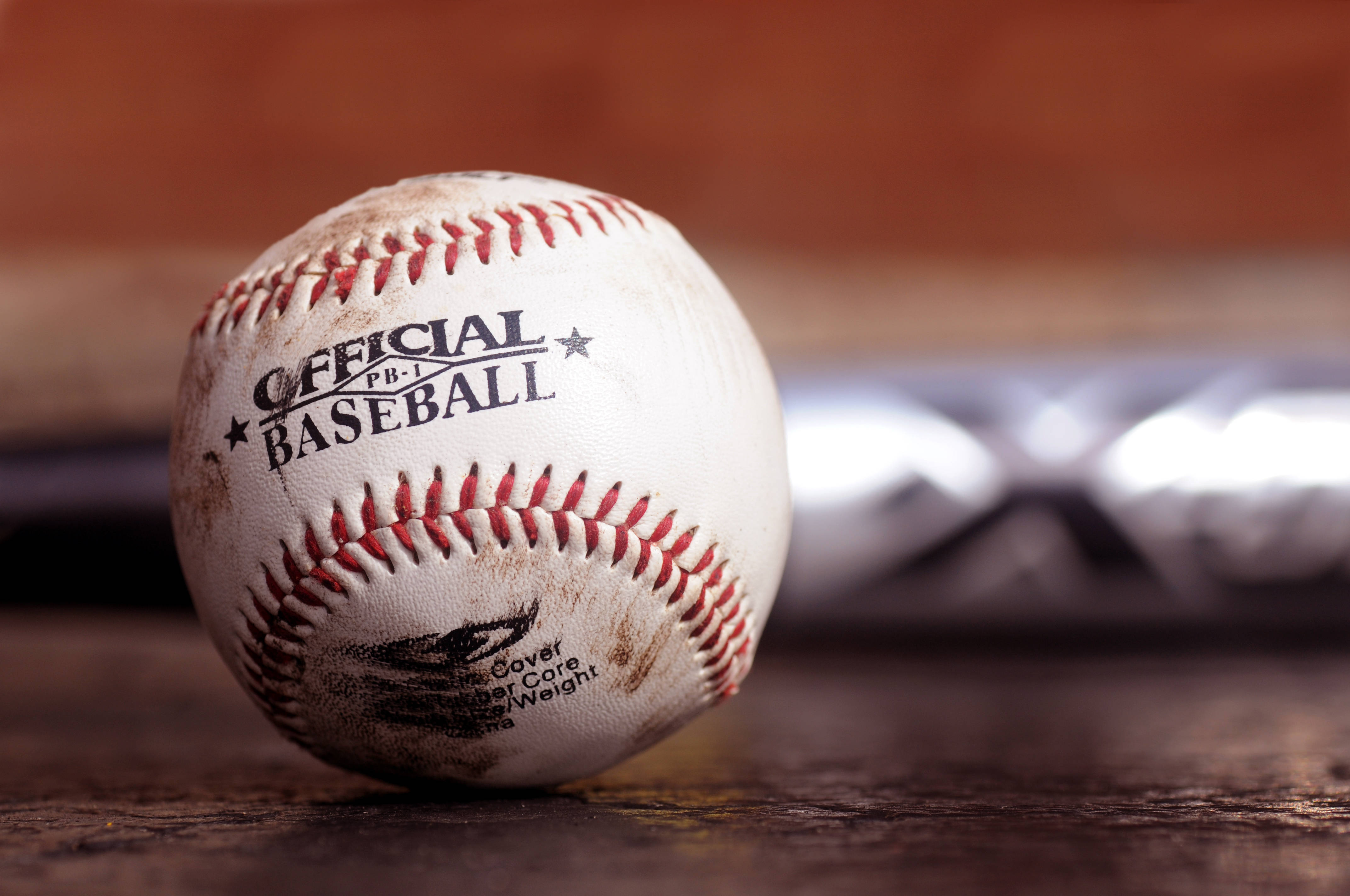 MLB 桌布 壁紙 — 一個磨損的官方棒球，帶有紅色縫線，放置在木質表面上，背景有模糊的球棒。