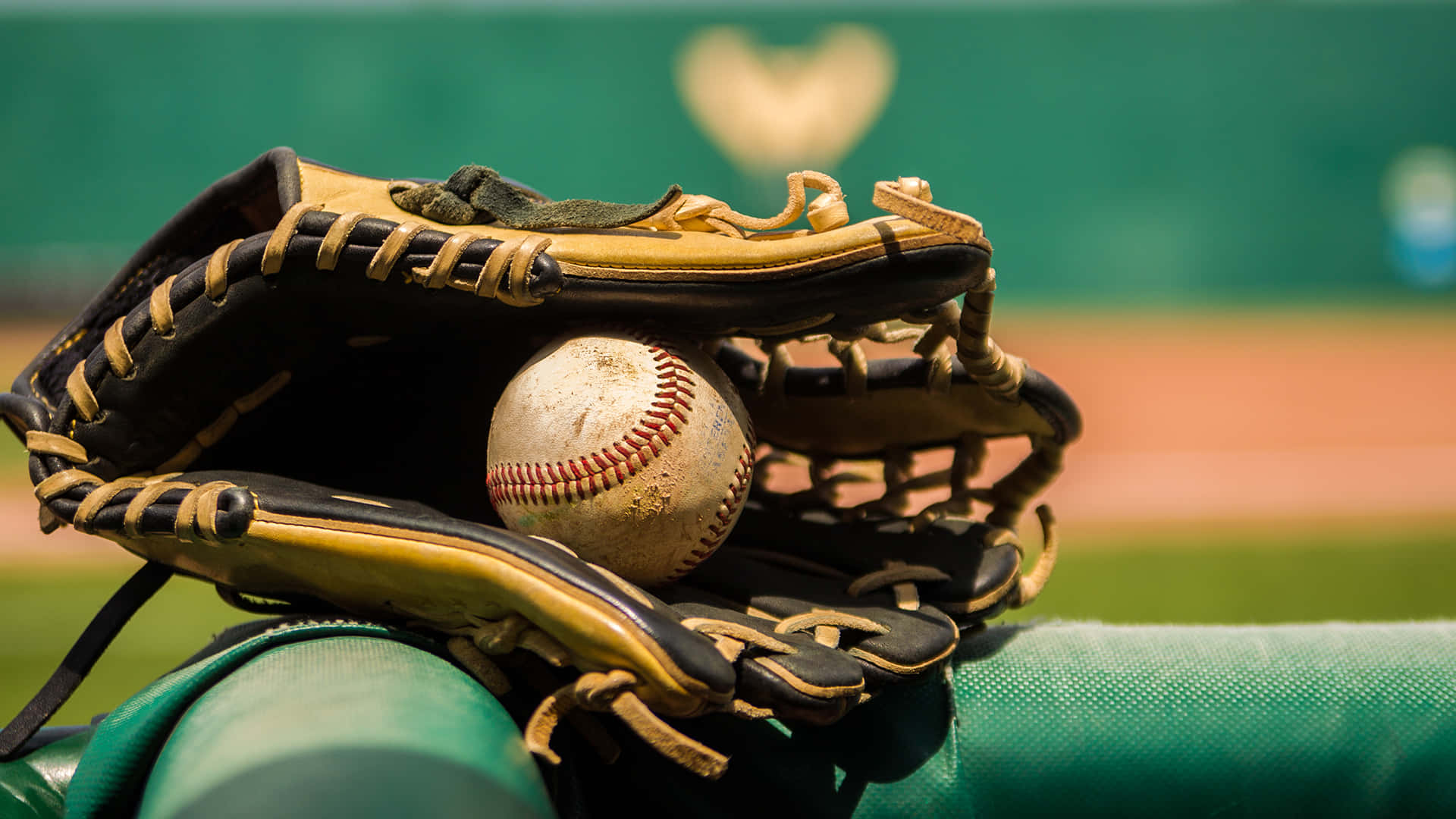 Baseball Gloves 桌布 壁紙 — 一個磨損的棒球手套，在模糊的綠色球場背景下，懷抱著一個有刮痕的棒球。