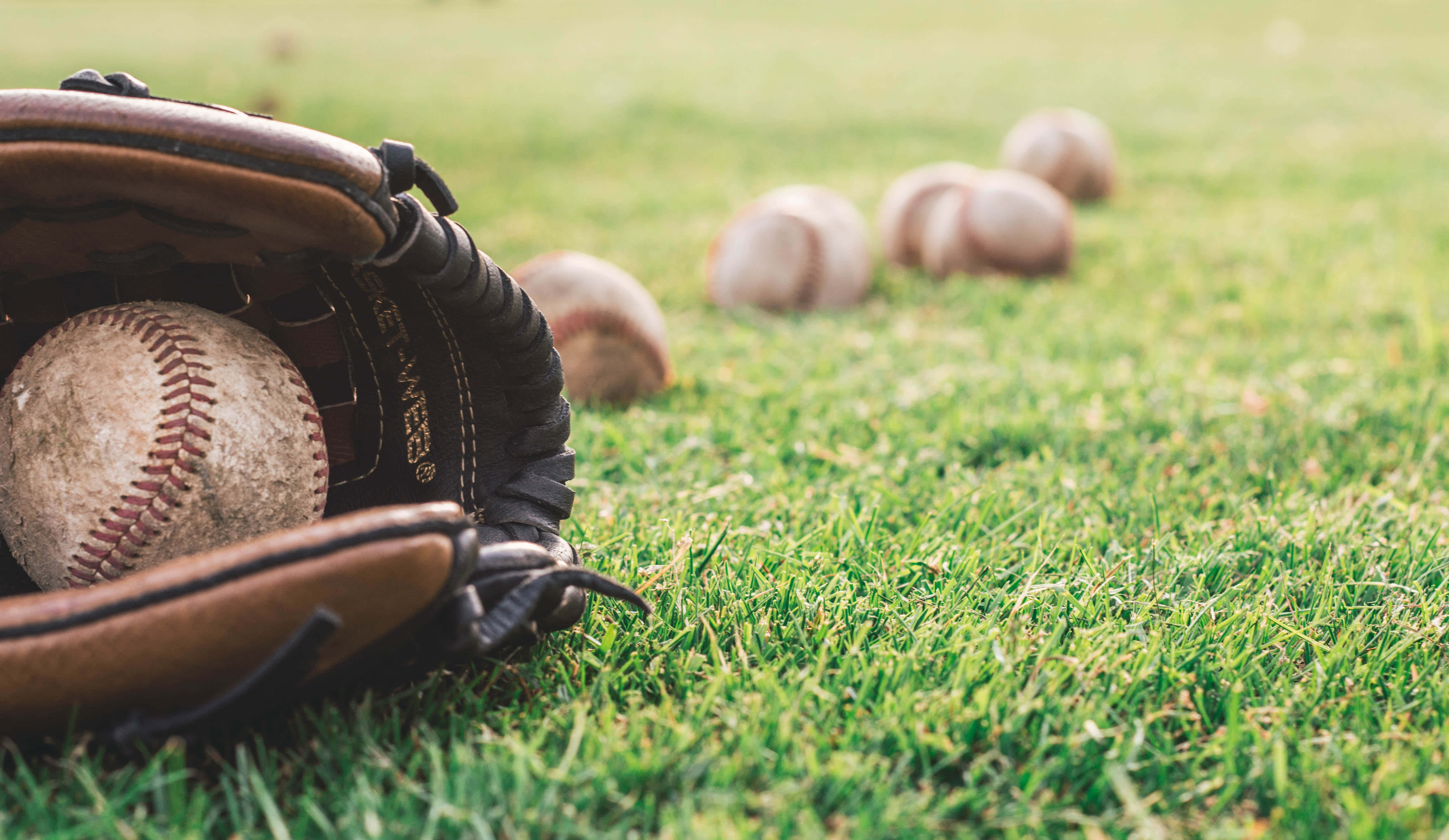 Baseball Gloves 桌布 壁紙 — 一個磨損的棒球手套在鮮綠色草地上托著一個用過的球，後面還有更多球以柔和焦點散落。