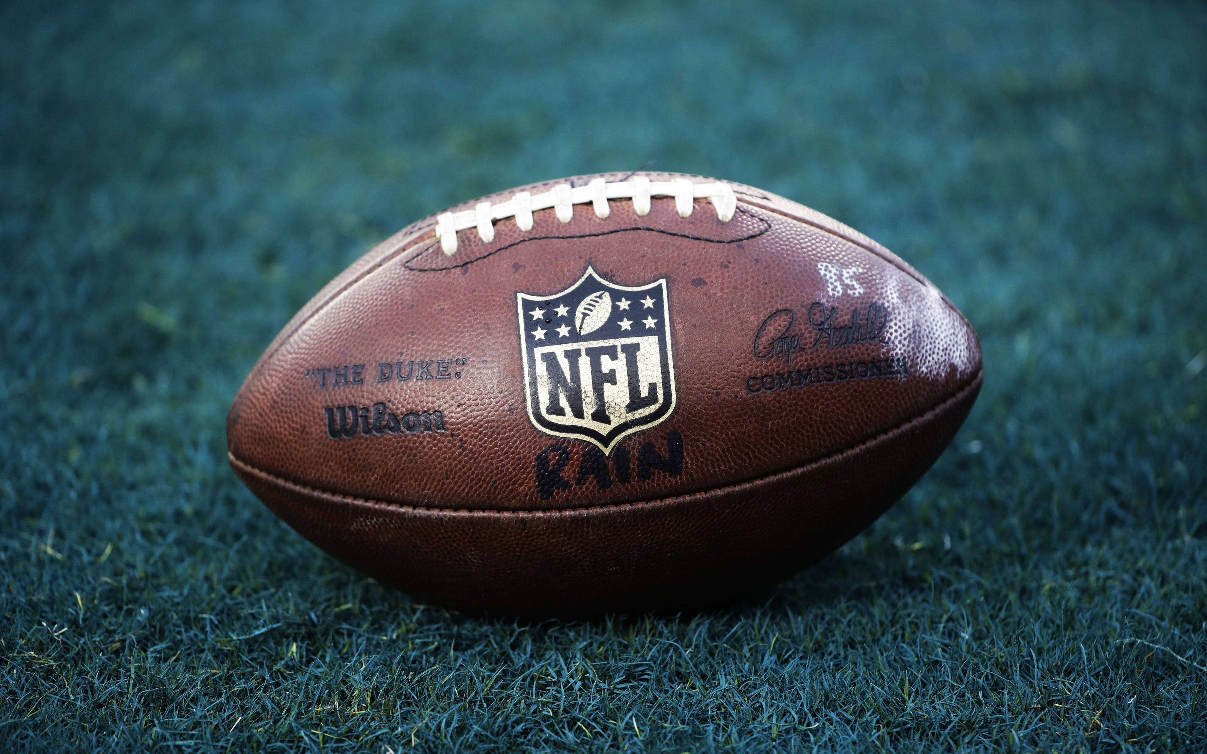 運動 桌布 壁紙 — 一個磨損的棕色NFL橄欖球在柔和光線下放在充滿活力的綠色草地上。