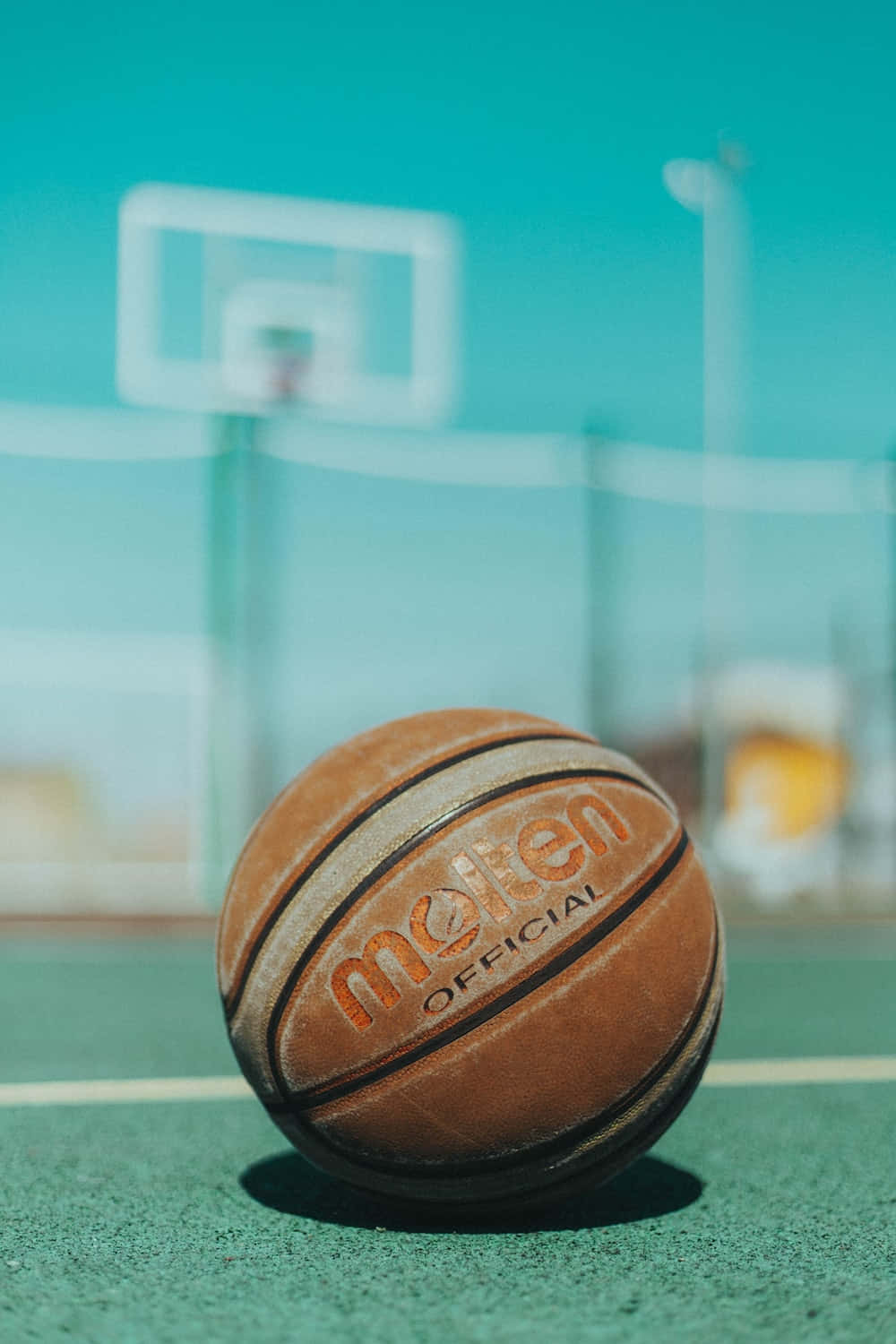 ball 桌布 壁紙 — 一個磨損的棕色籃球放在綠色球場上，背景有模糊的籃框和藍綠色天空。