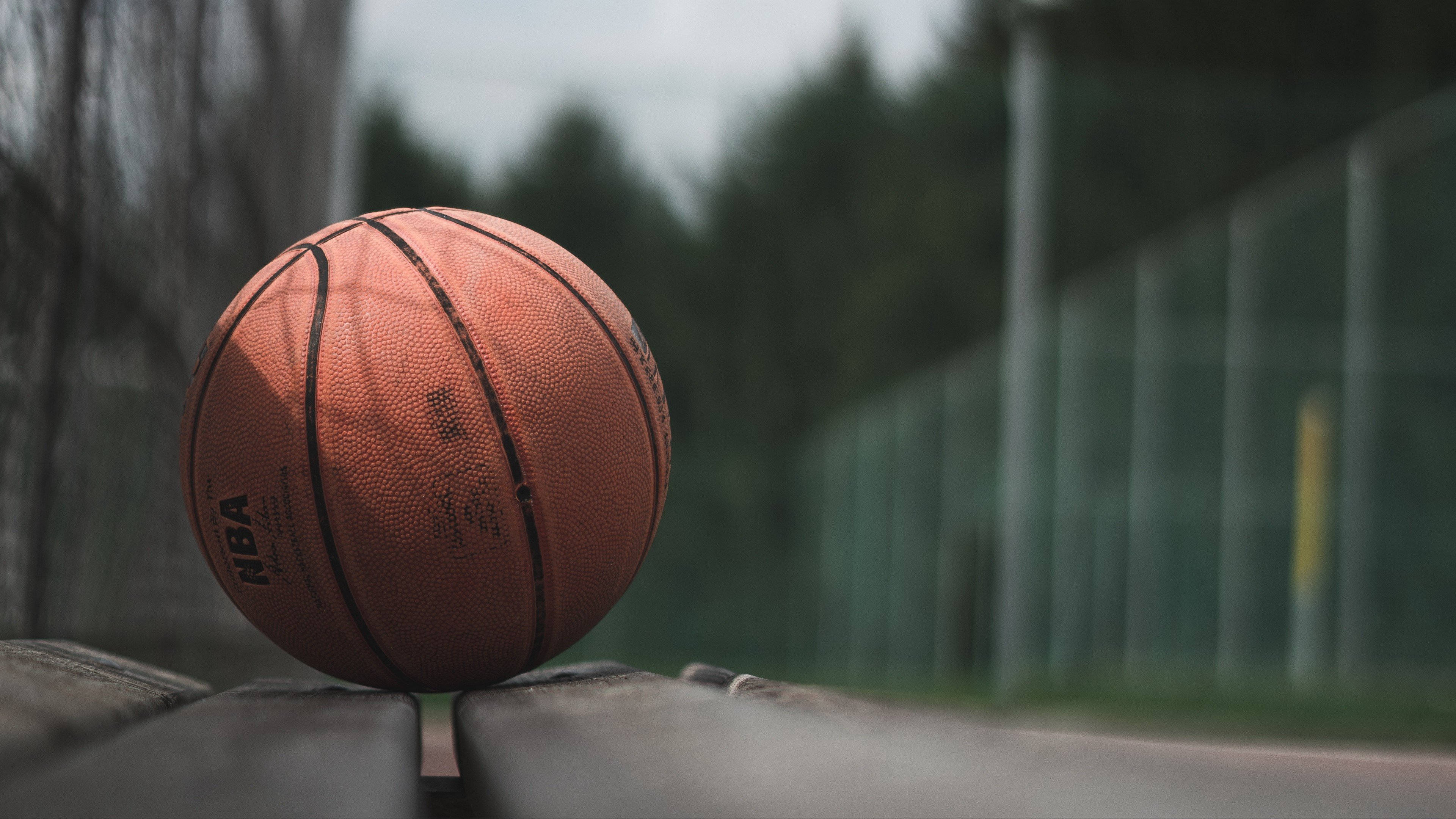 4K籃球壁紙 桌布 壁紙 — 一個磨損的橙色NBA籃球放在長椅上，背景是模糊的戶外球場和樹木。