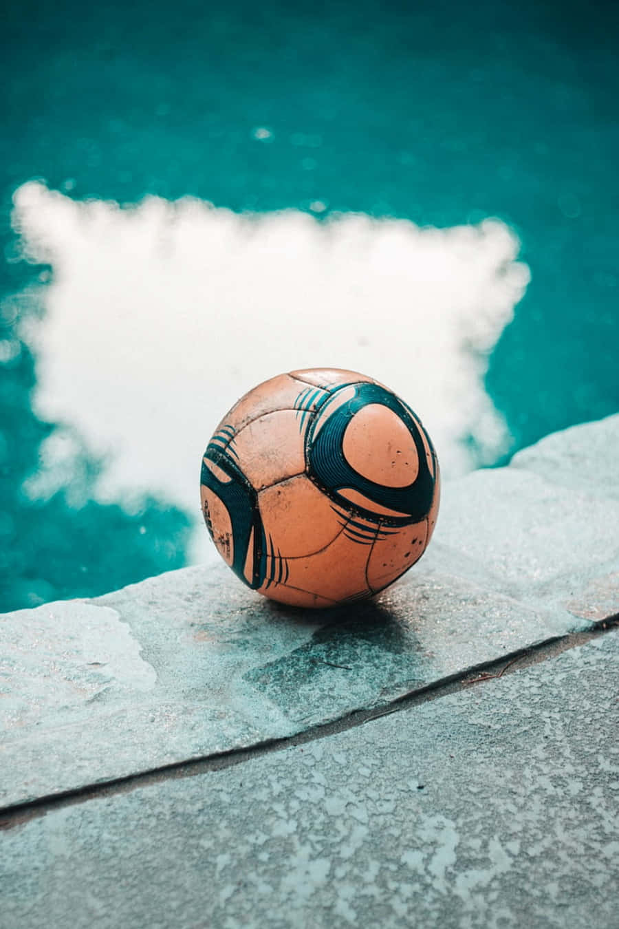 Soccer Ball 桌布 壁紙 — 一個磨損的橙色和黑色足球放在濕混凝土泳池邊緣，旁邊是藍綠色的水。