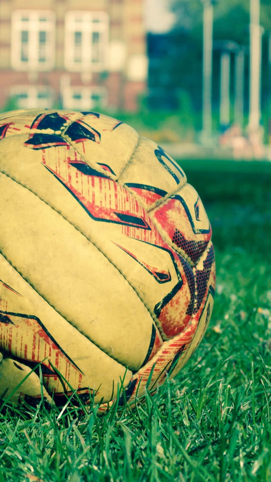 Soccer iPhone 桌布 壁紙 — 一個磨損的足球，帶有紅色、黑色和黃色圖案，放在鮮綠色的草地上，背景是一座模糊的磚砌建築。