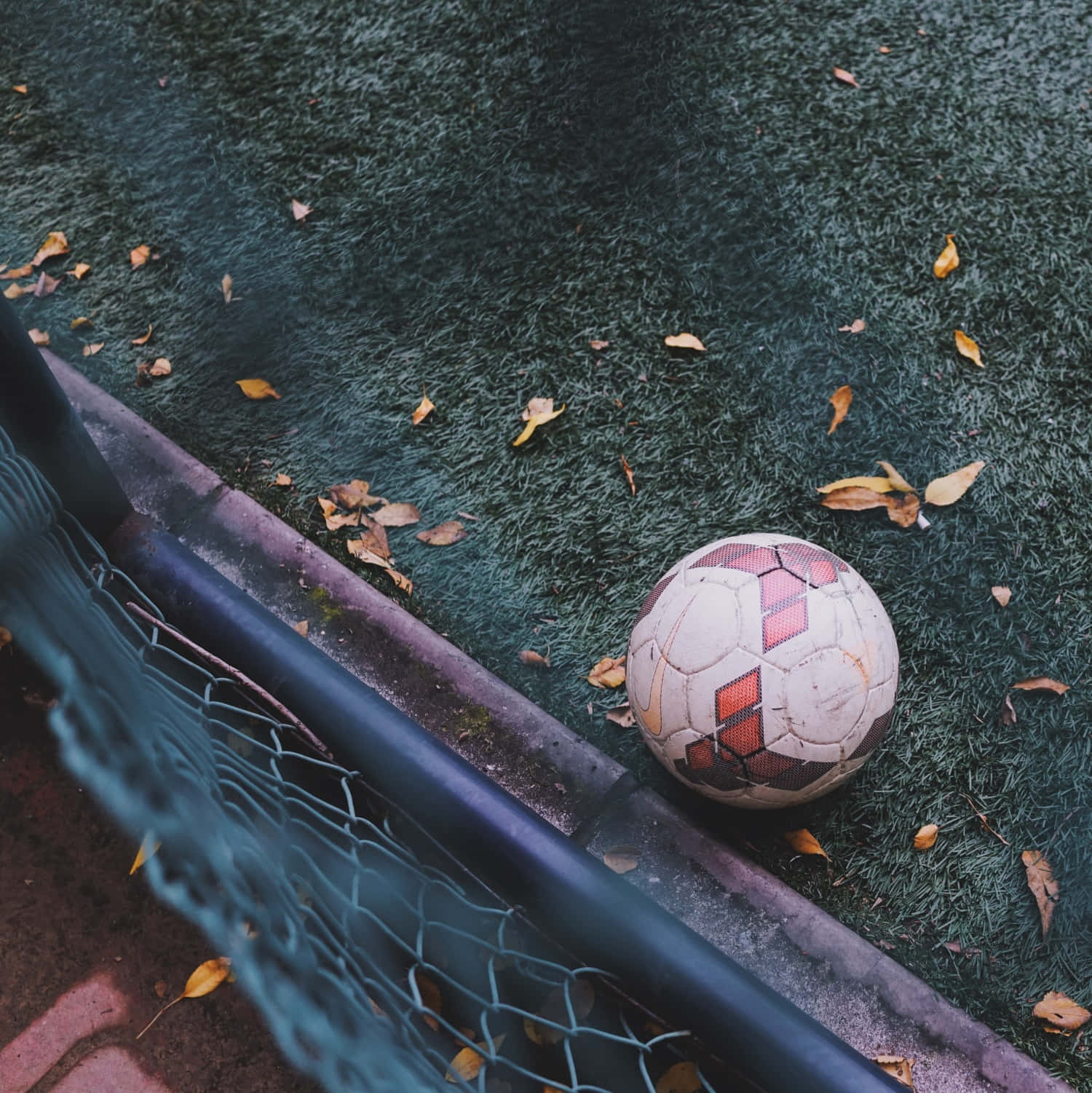 Soccer Ball 桌布 壁紙 — 一個磨損的足球放在綠色人造草皮上，旁邊的球門網散落著秋天的落葉。