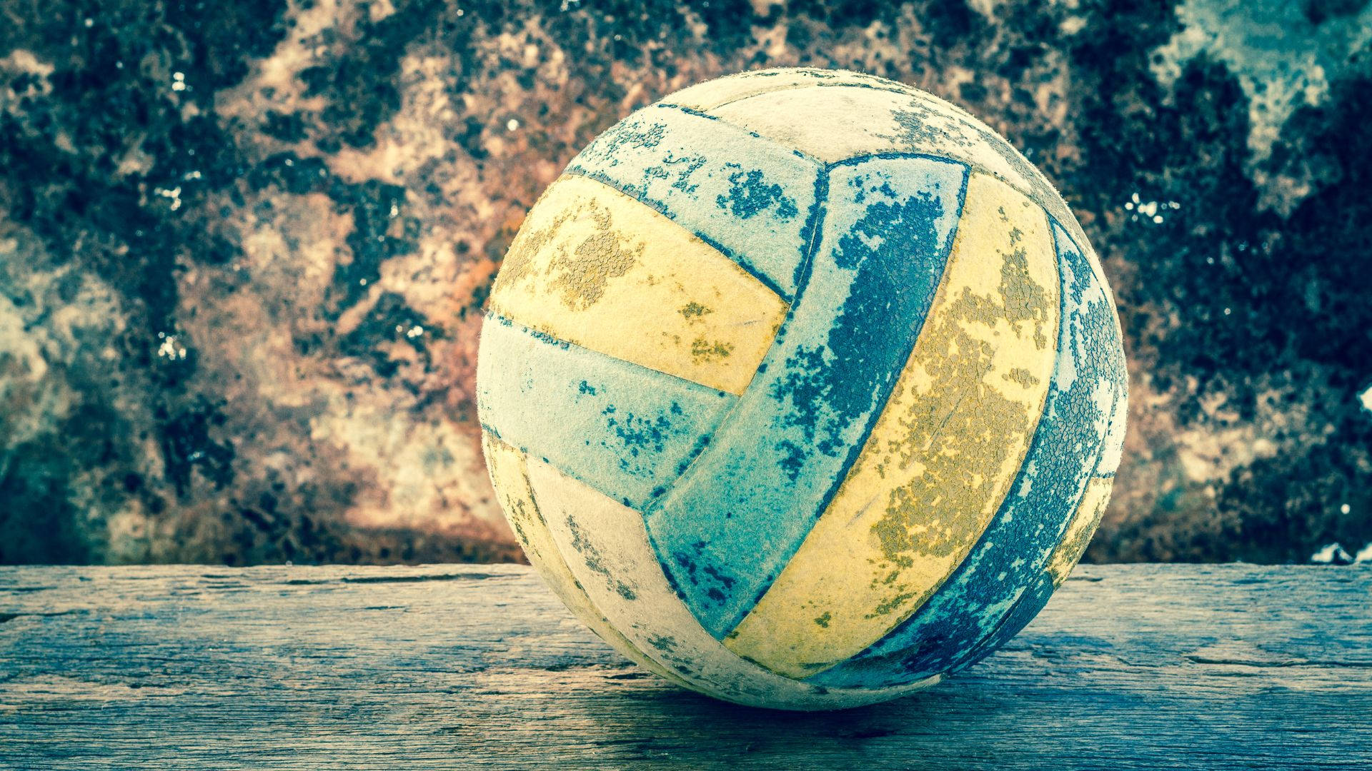 Volleyball Aesthetic 桌布 壁紙 — 一個磨損的黃藍色排球放在經風化的木質表面上，背景是紋理石牆。