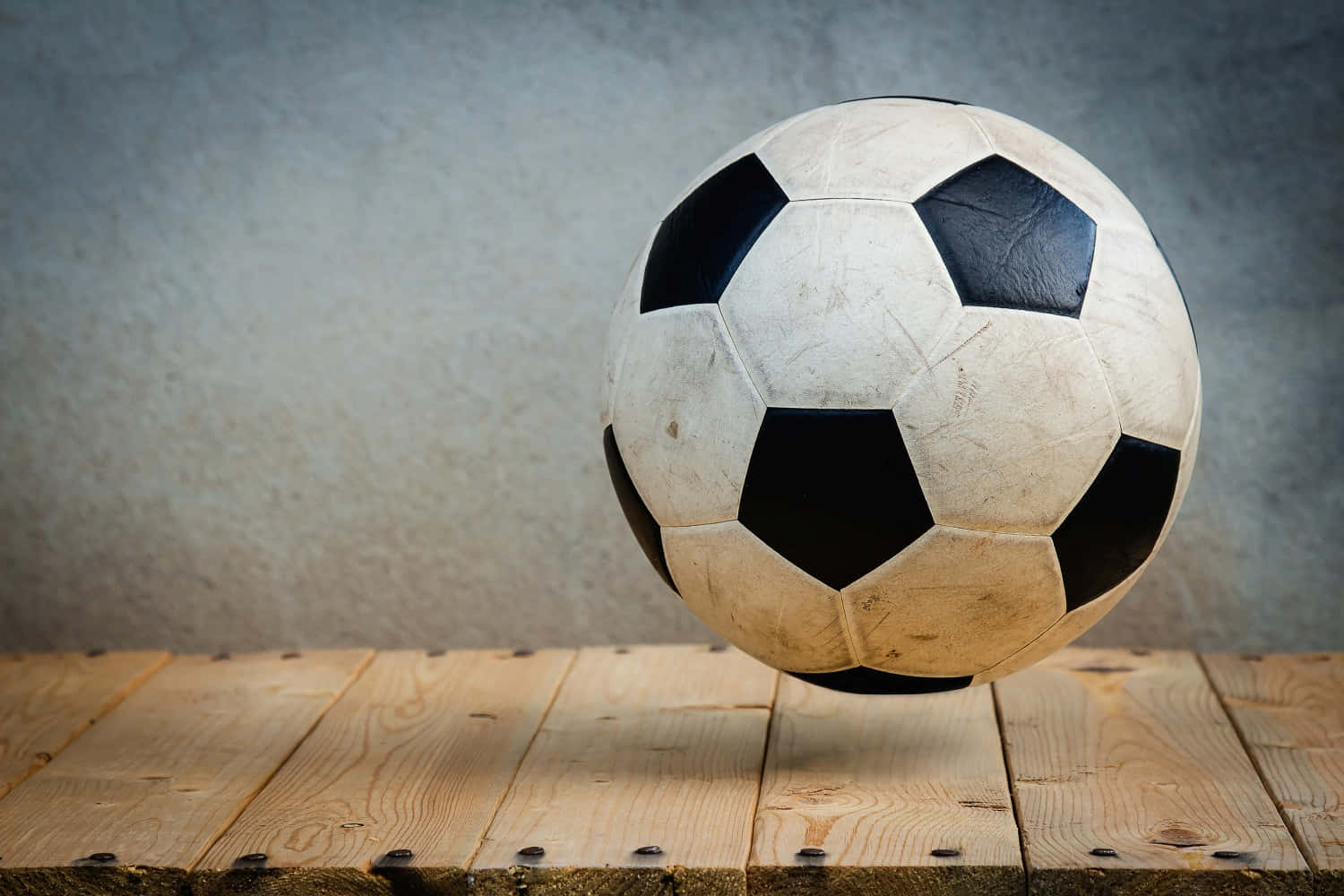 Soccer Ball 桌布 壁紙 — 一個磨損的黑白足球放在質樸的木質表面上，背景是帶紋理的灰色牆壁。