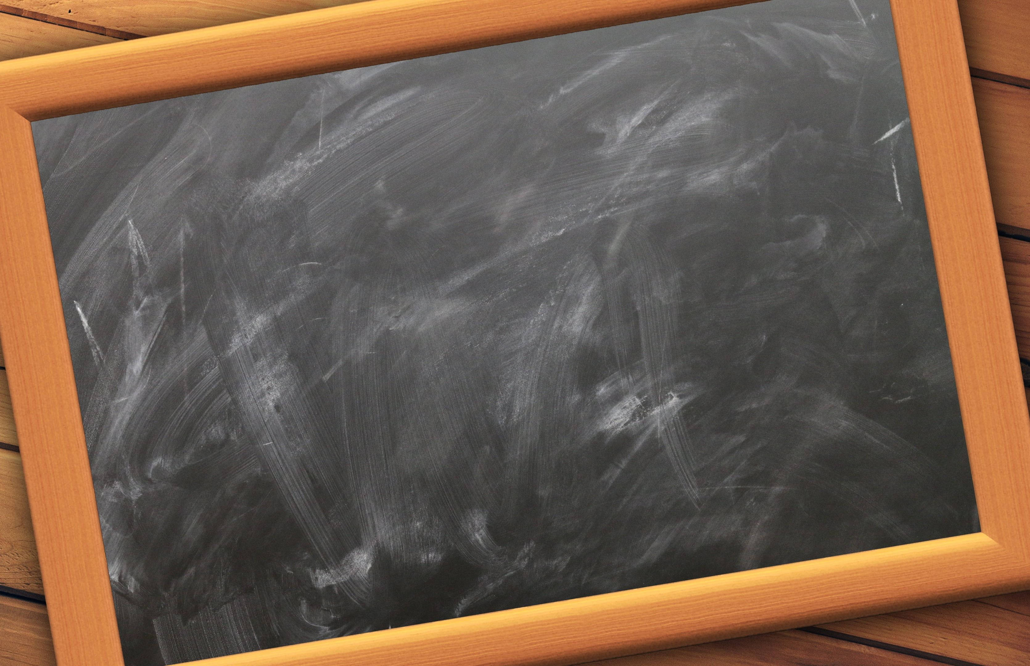 Chalkboard 桌布 壁紙 — 一個磨損的黑色黑板特寫，帶有白色粉筆污跡，鑲嵌在淺色木框中，放置在木質表面上。