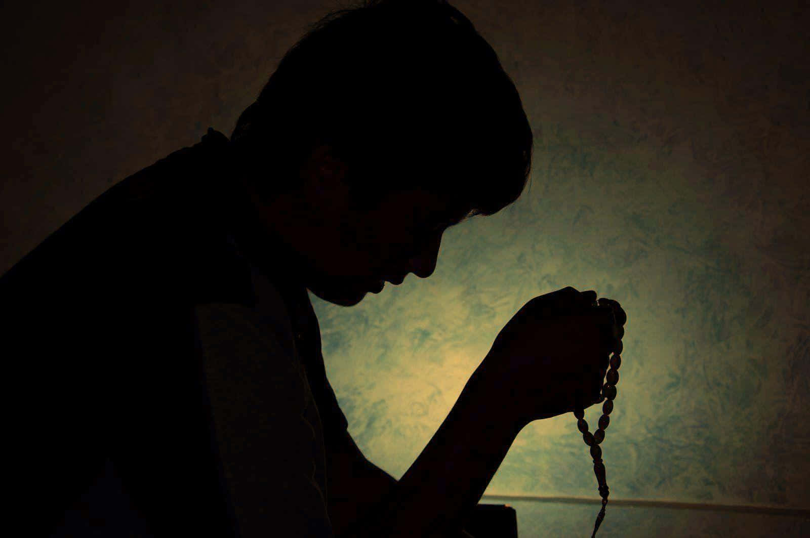 Praying 桌布 壁紙 — 一個祈禱的人的剪影，握著玫瑰經念珠，靠著一面有紋理的牆，被溫暖的背光包圍。