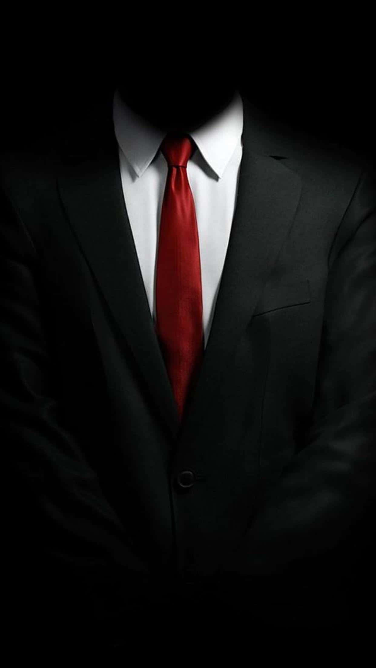iPhone X Hitman 2 桌布 壁紙 — 一個神秘人物身穿黑色西裝和紅色領帶，站在黑暗中，臉部被陰影遮蓋。
