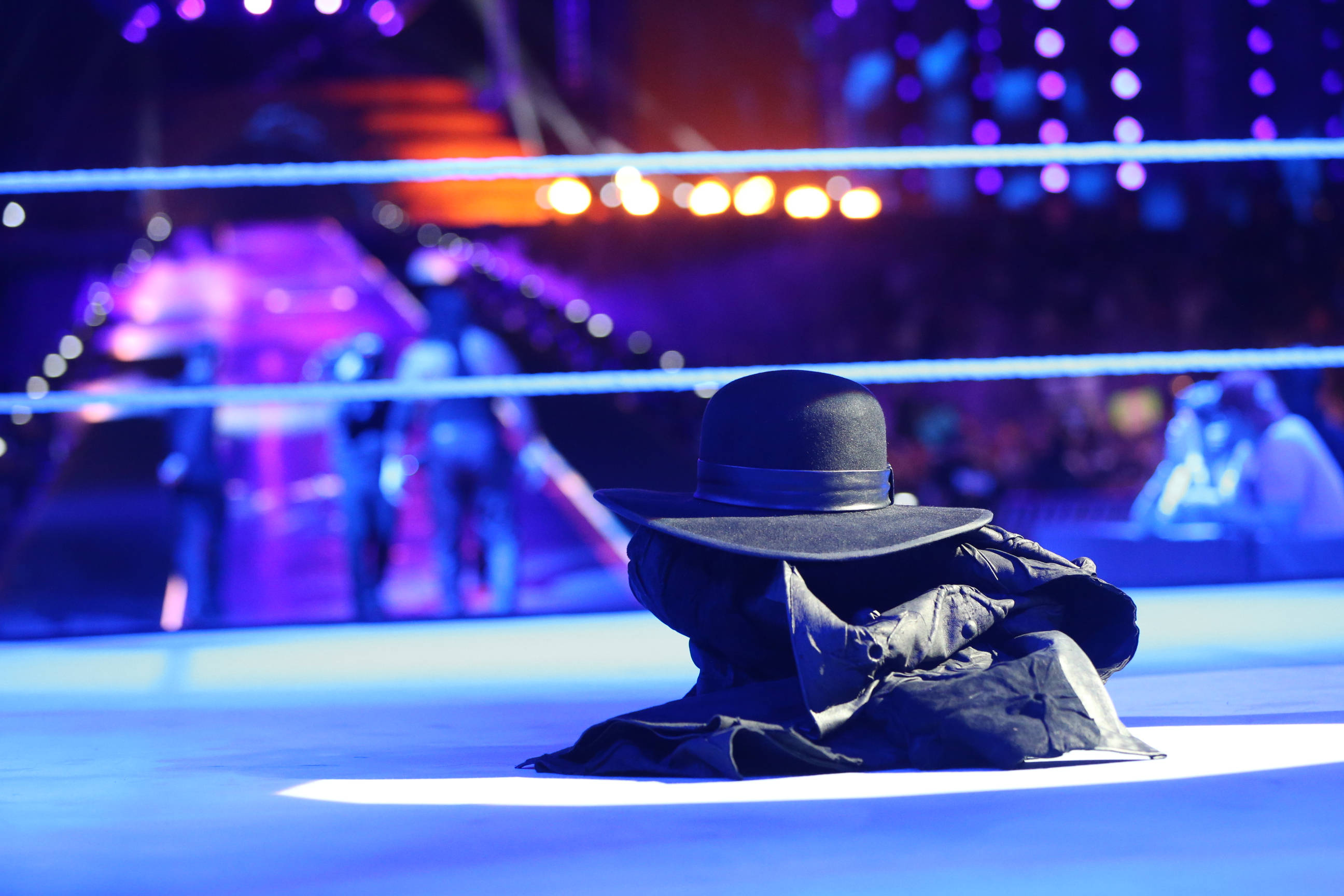 Undertaker 桌布 壁紙 — 一個神秘的摔角手戴著黑色帽子和外套，獨自坐在明亮的摔角擂台中，紫色舞台燈光下。