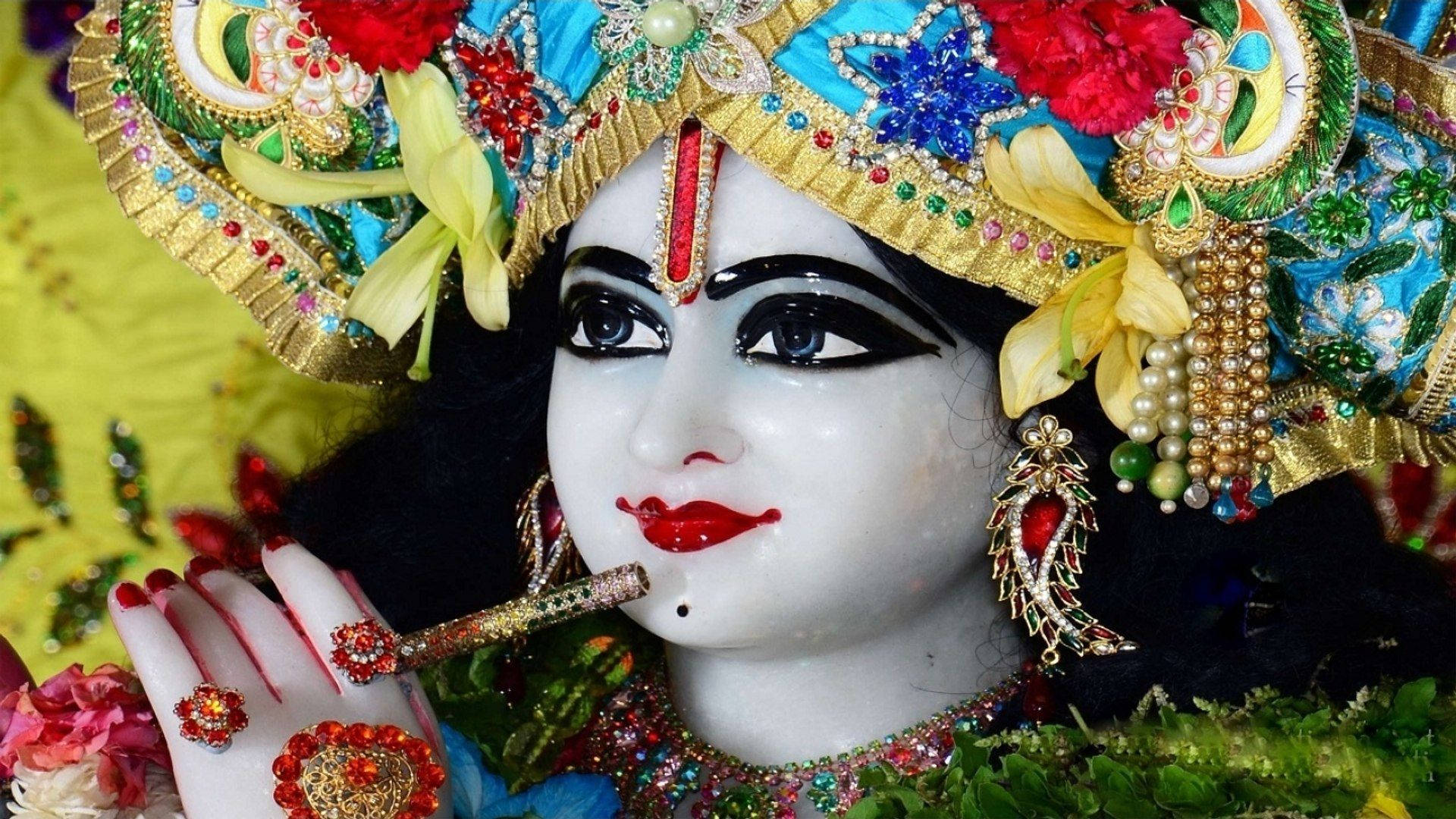krishna god 桌布 壁紙 — 一個神聖偶像的特寫，裝飾著鮮豔的寶石、花朵和精緻的金色頭飾，將笛子放在唇邊。