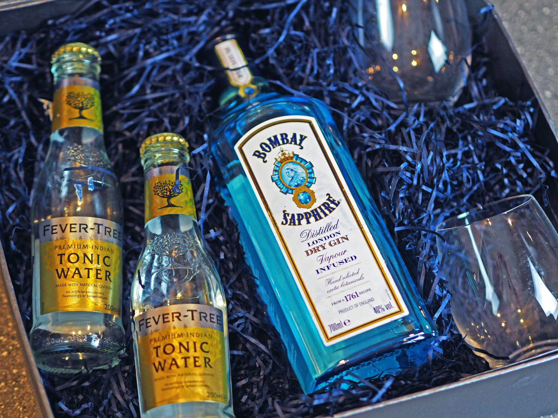  壁紙 — 一個禮物盒，內有Bombay Sapphire琴酒、兩瓶Fever Tree通寧水、兩個酒杯，放在藍色碎紙中。