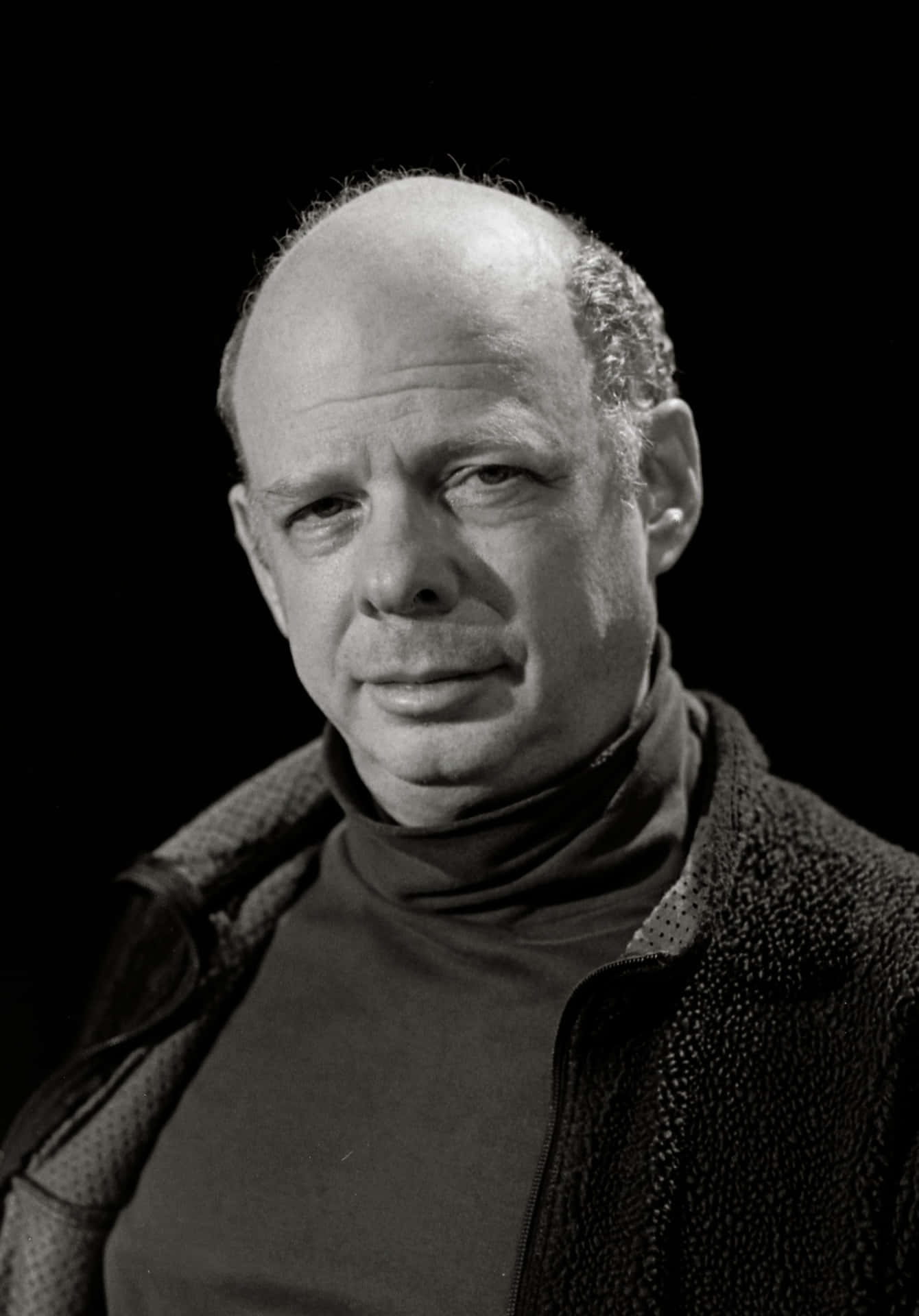 Wallace Shawn 桌布 壁紙 — 一個禿頭男子穿著高領毛衣和質感外套，表情嚴肅，在深色背景前的黑白肖像。