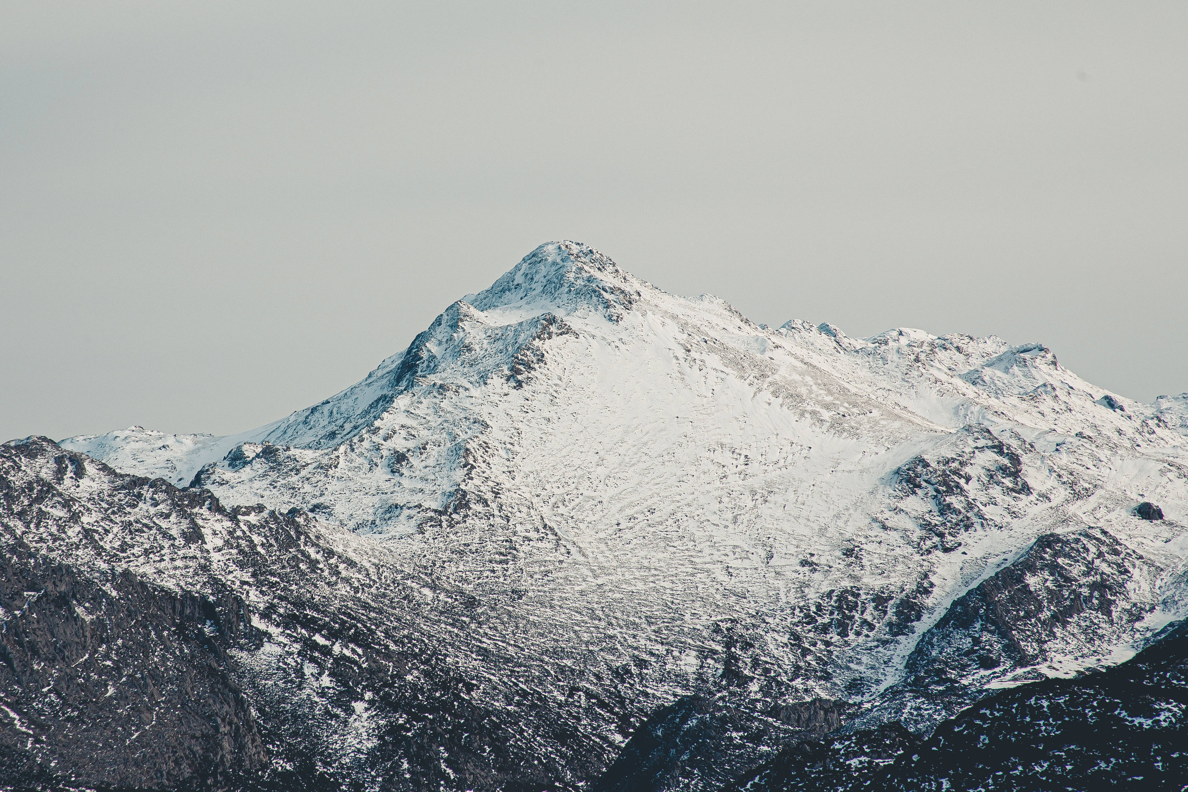 Most Beautiful Nature 桌布 壁紙 — 一個積雪的山峰在淡灰色的天空下陡峭地聳立，下方是質感豐富的岩石斜坡。