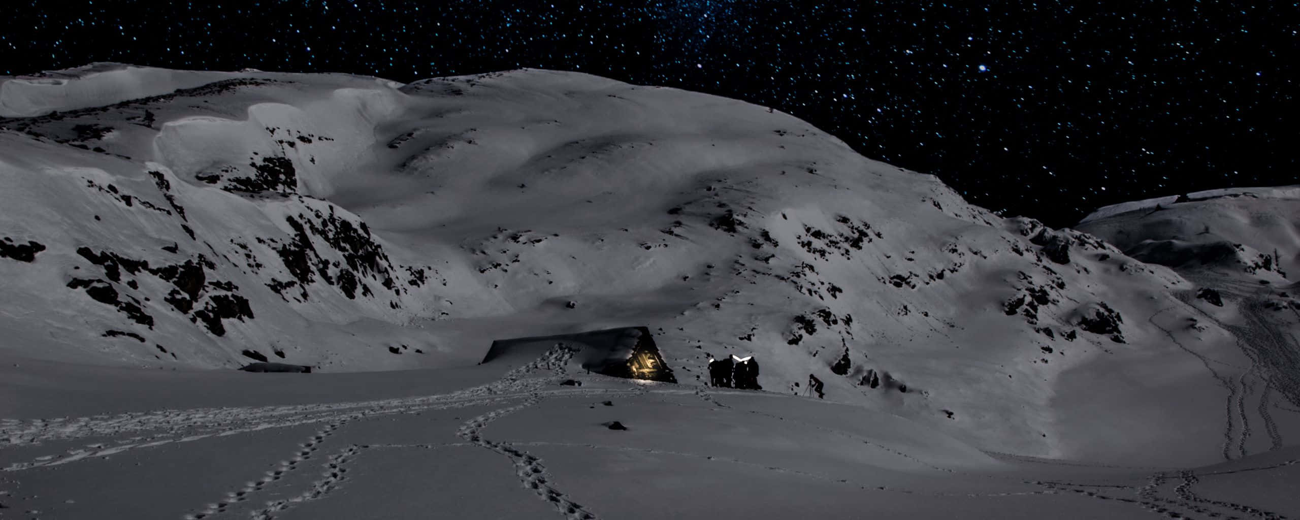 山景 桌布 壁紙 — 一個積雪的山洞在星空下溫暖地發光，腳印通向洞口。