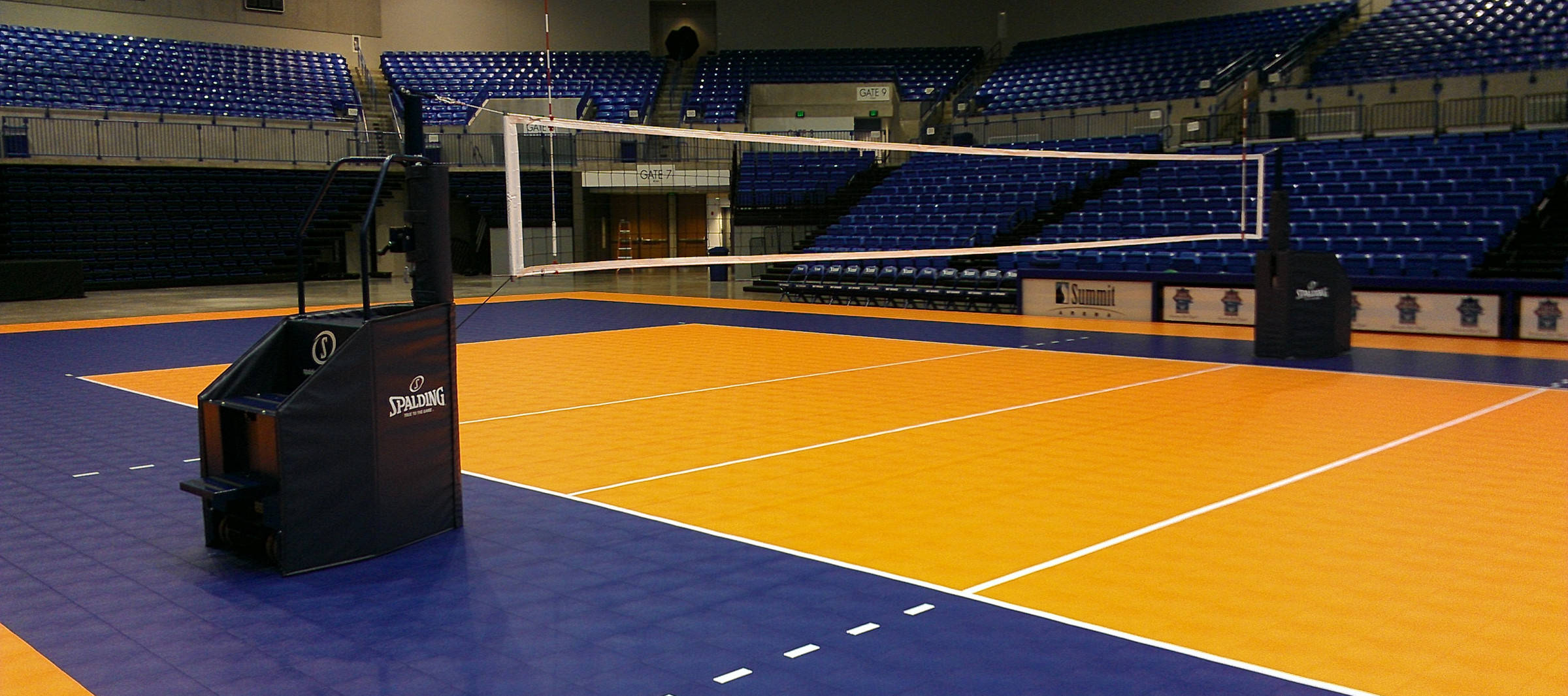 Volleyball 桌布 壁紙 — 一個空曠的室內排球場，設有藍色座位和橙色比賽場地，在明亮的人造燈光下。