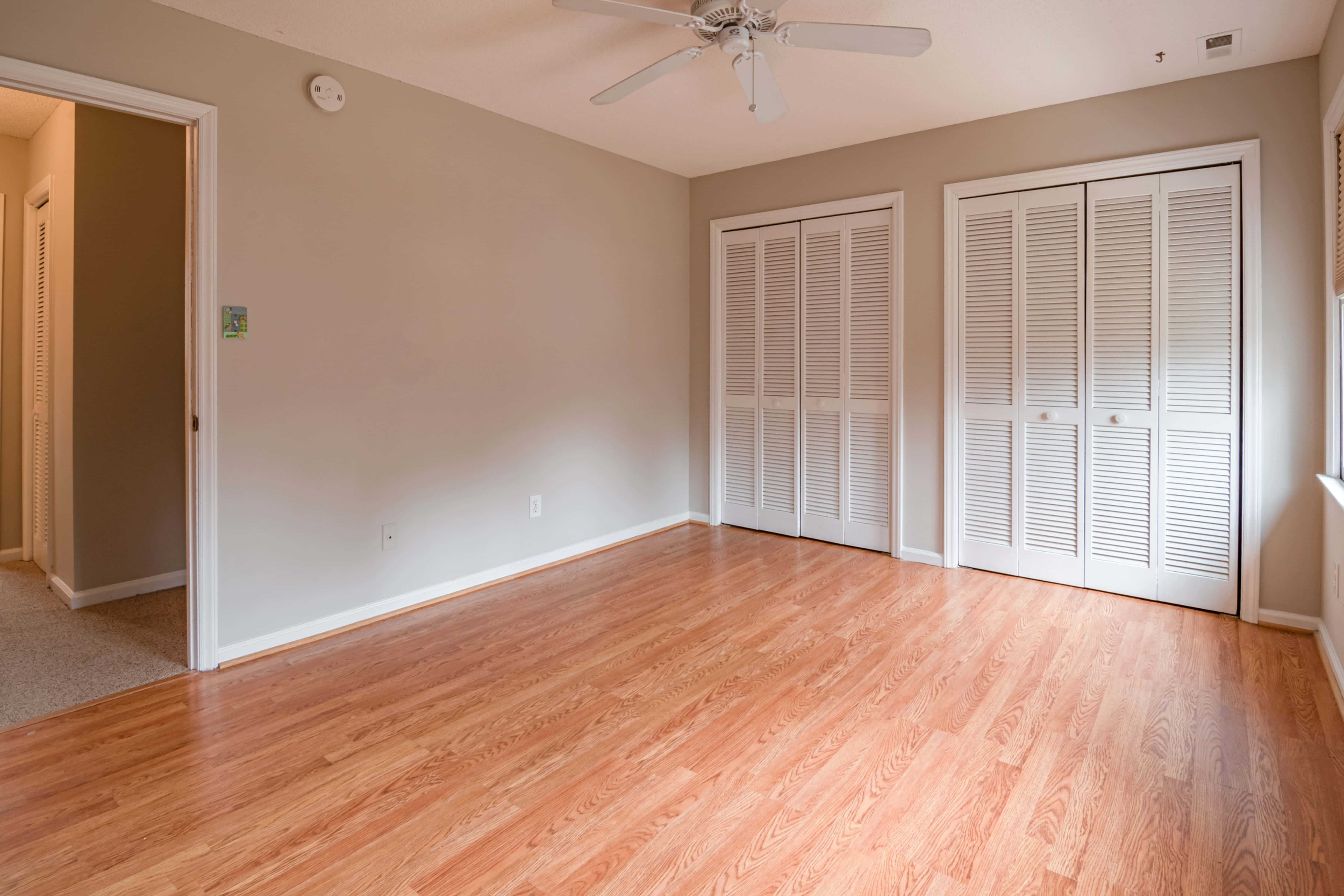 Empty Room 桌布 壁紙 — 一個空置的臥室，舖有硬木地板、米色牆壁和白色百葉衣櫃門，天花板上裝有吊扇。