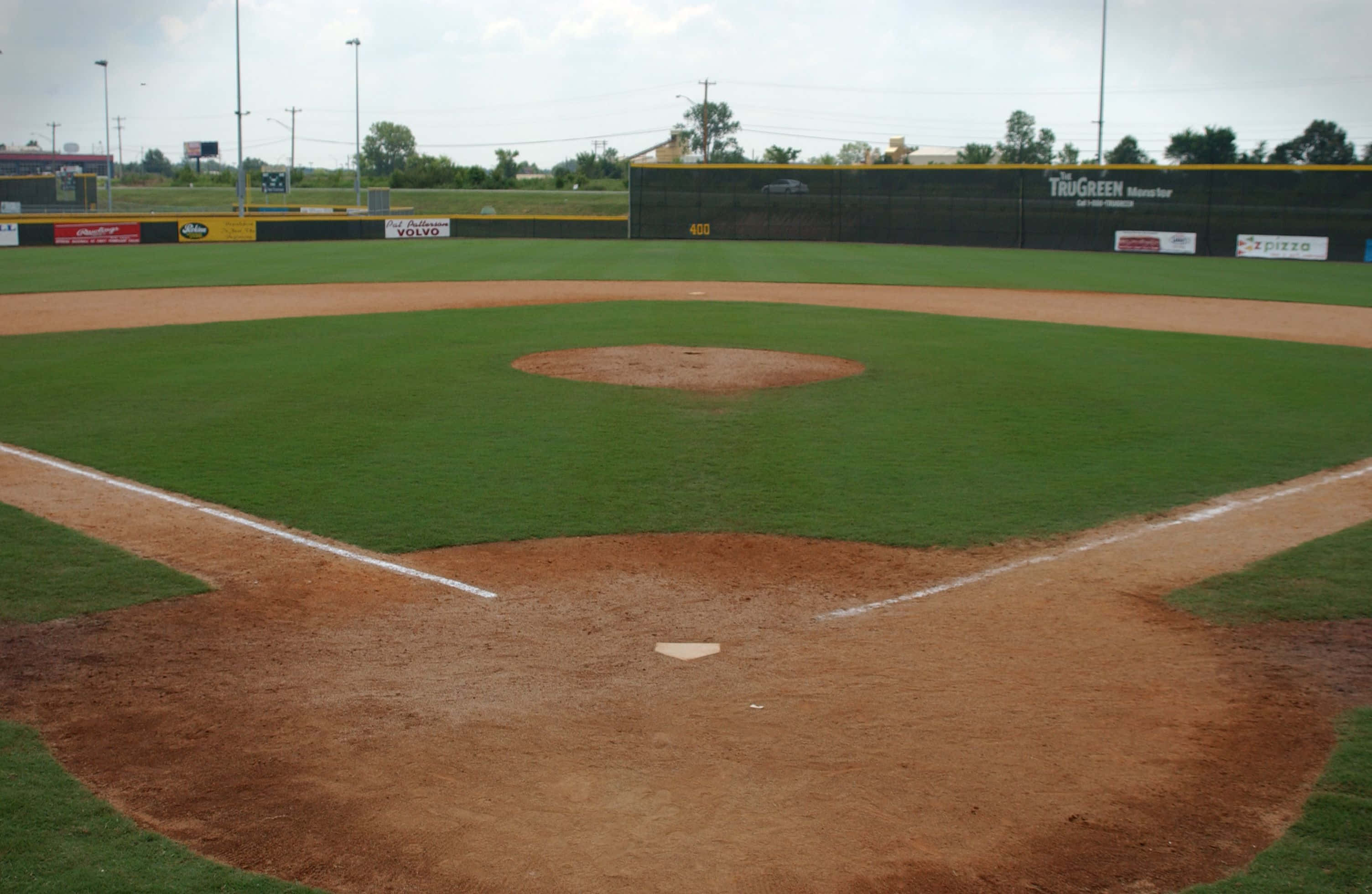 Baseball Field 桌布 壁紙 — 一個空蕩蕩的棒球場，有充滿活力的綠色草地和棕色泥土，天空多雲。