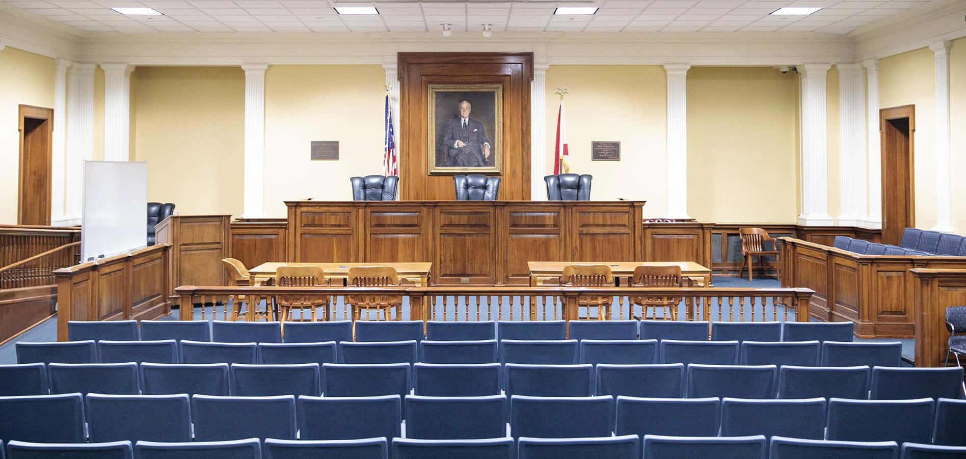 Courtroom 桌布 壁紙 — 一個空蕩蕩的法庭，設有木製長椅、藍色觀眾席，以及法學家畫像下的法官席。