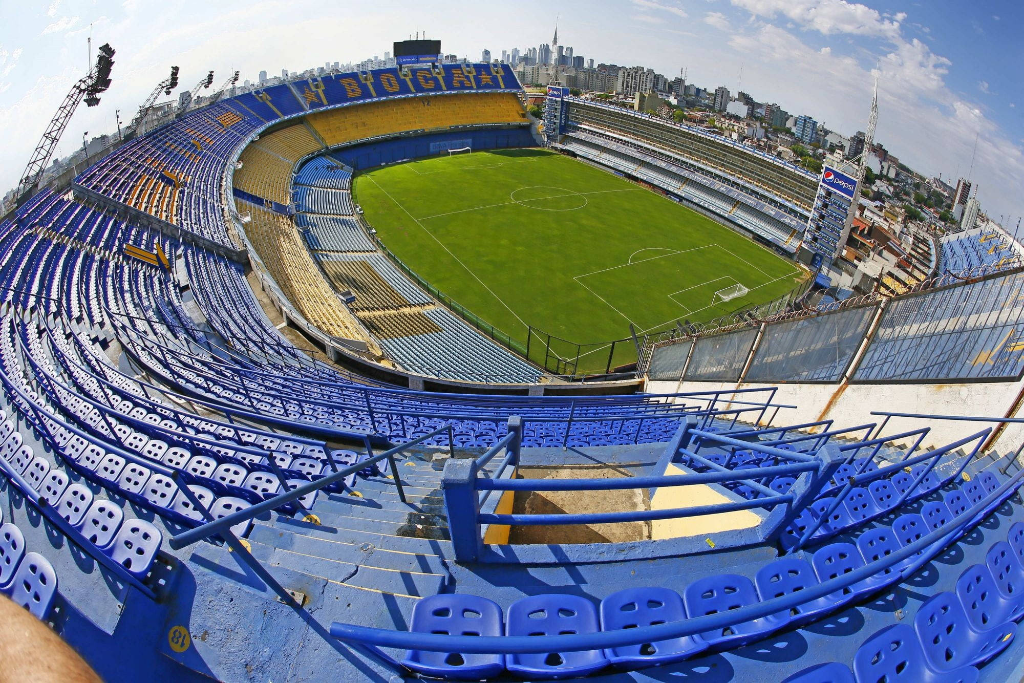 Stadium 桌布 壁紙 — 一個空蕩蕩的藍黃色足球場的魚眼視角，球場上鋪著充滿活力的綠色草坪，上方是晴朗明亮的天空。