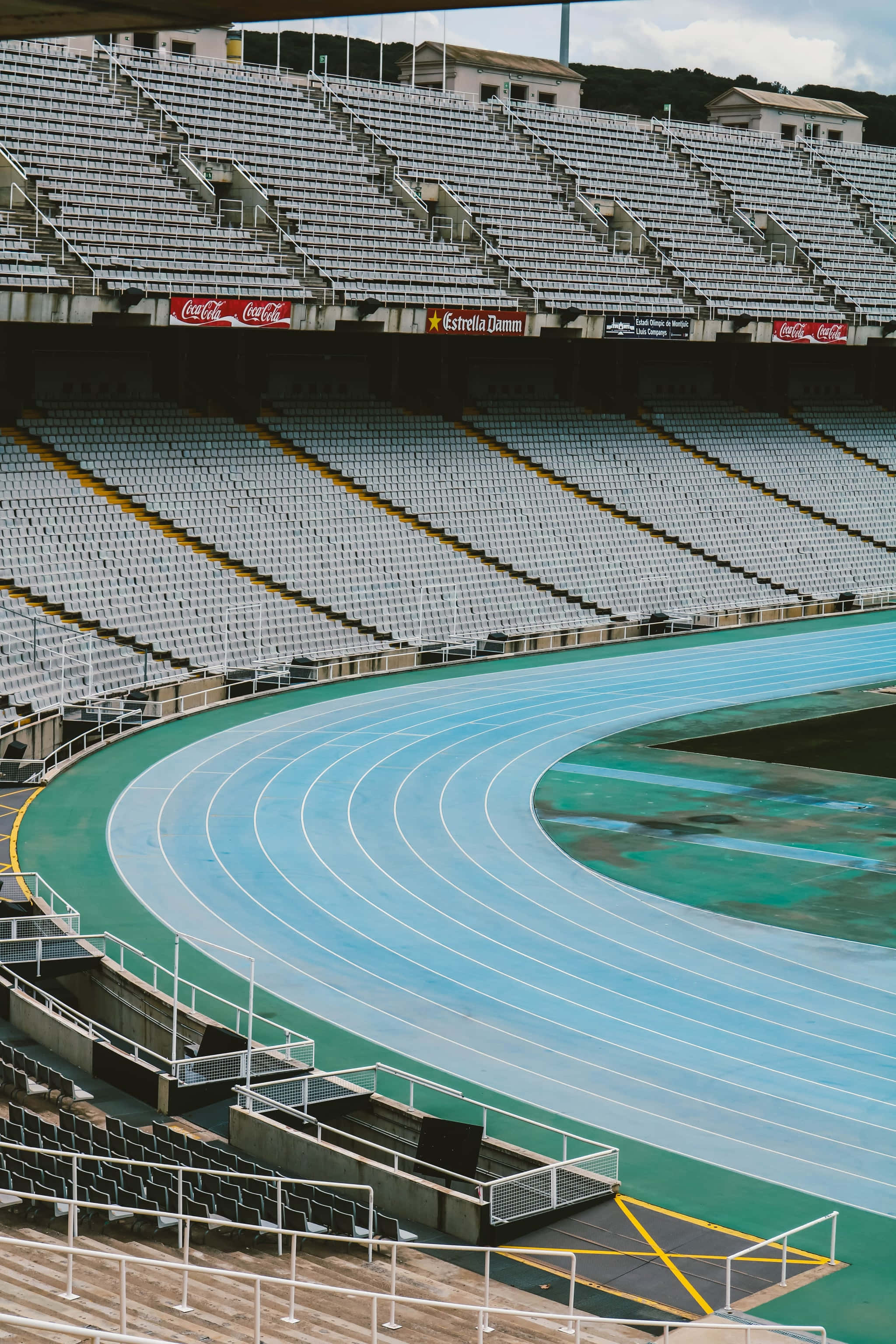 track 桌布 壁紙 — 一個空蕩蕩的體育場，設有藍色跑道和白色座位，天空多雲。